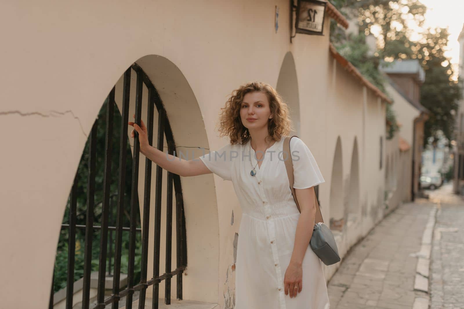 A woman with curly hair is holding on to a bar of a metal lattice in an old European town. A lady in a white dress in the evening.