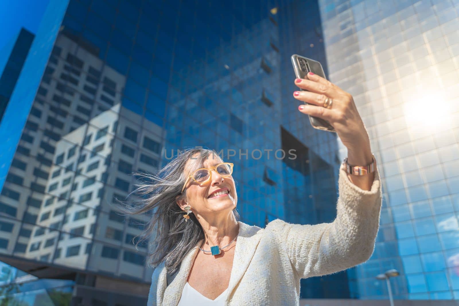 Happy businesswoman smiling and taking a selfie with mobile phone in front of business buildings. High quality photo