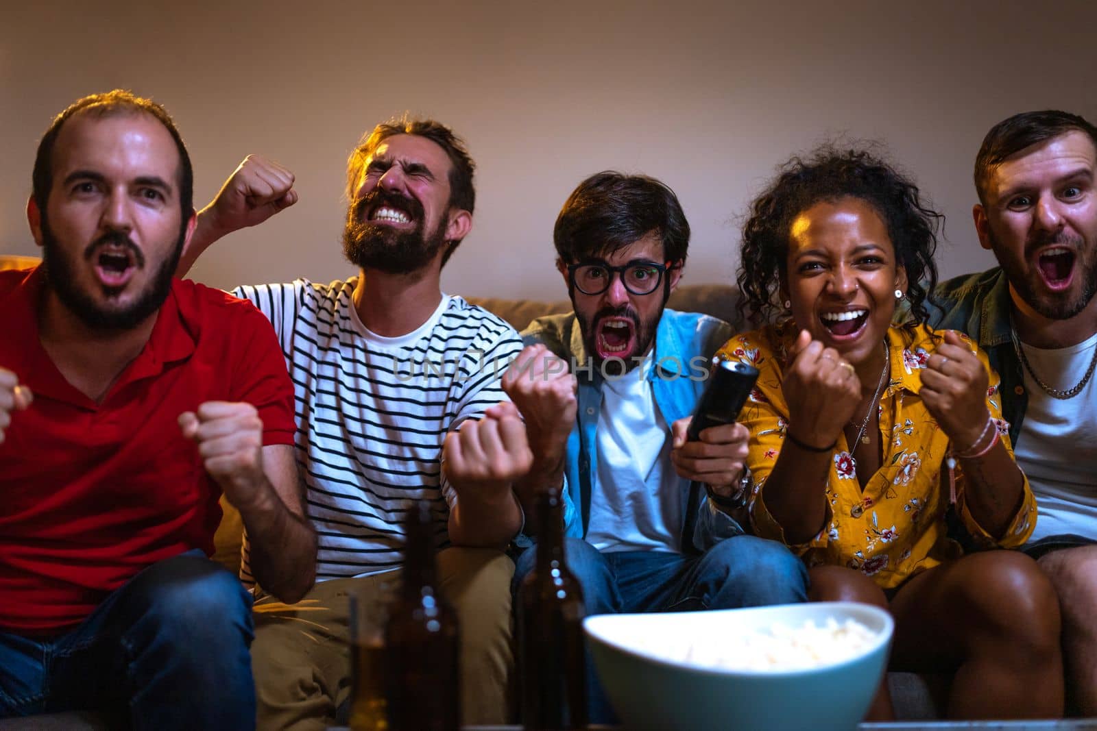Group of friends watching football game on tv celebrating teams scoring goal. Soccer. Looking at camera. by Hoverstock