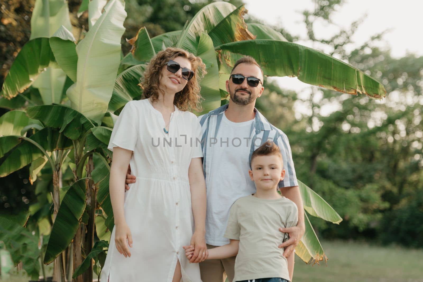Father, mother, and son are posing with palm trees in the background. A close photo of a happy family in the garden in summer at sunset.