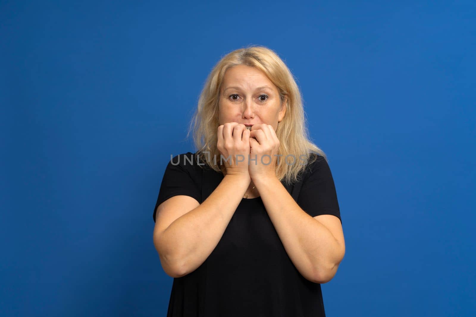 Caucasian blonde woman biting her nails out of fear, she is experiencing a dangerous and terrifying situation. Isolated on a blue studio background.