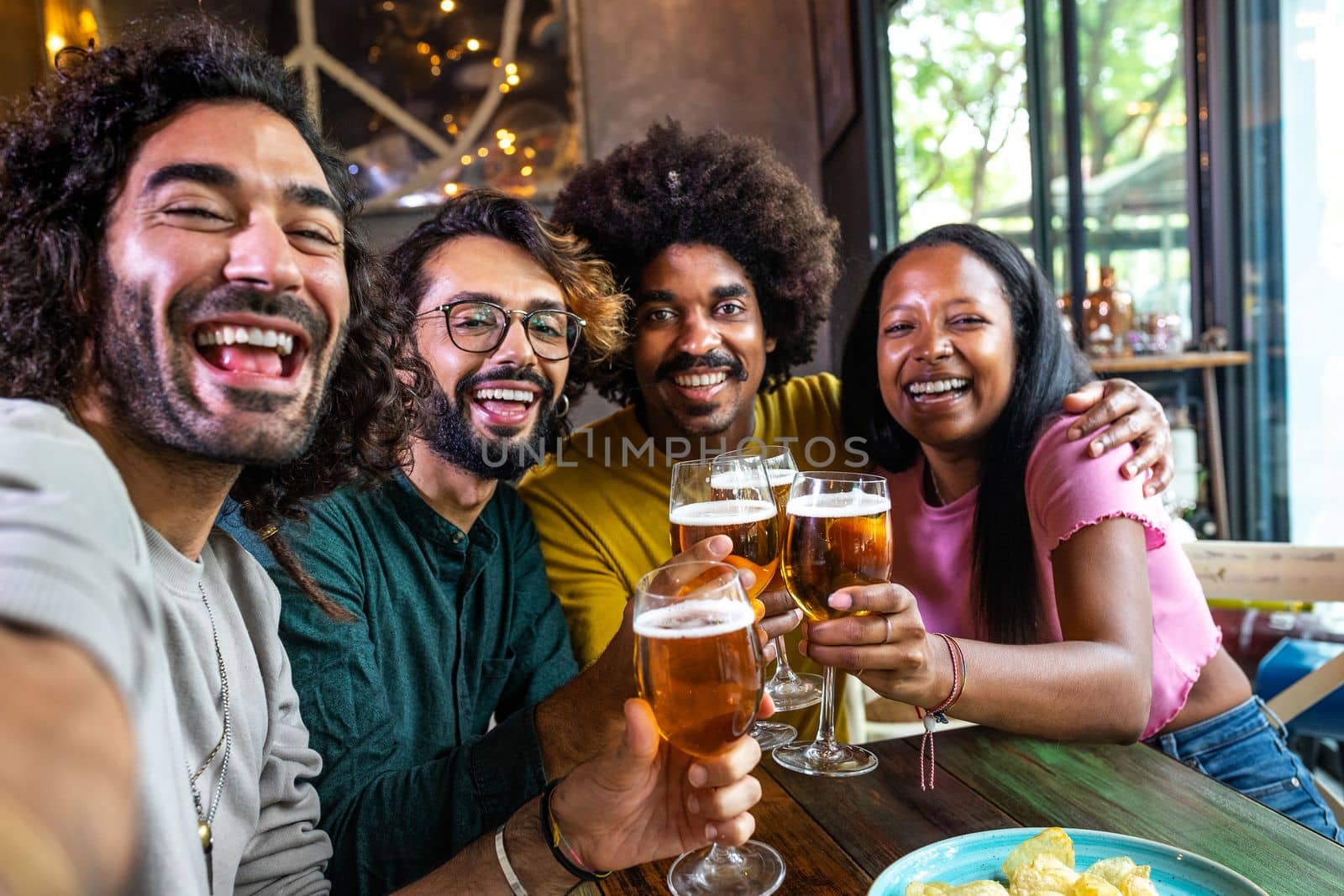 Happy multiracial friends in a bar toasting with beer taking selfie with phone looking at camera. Lifestyle concept.