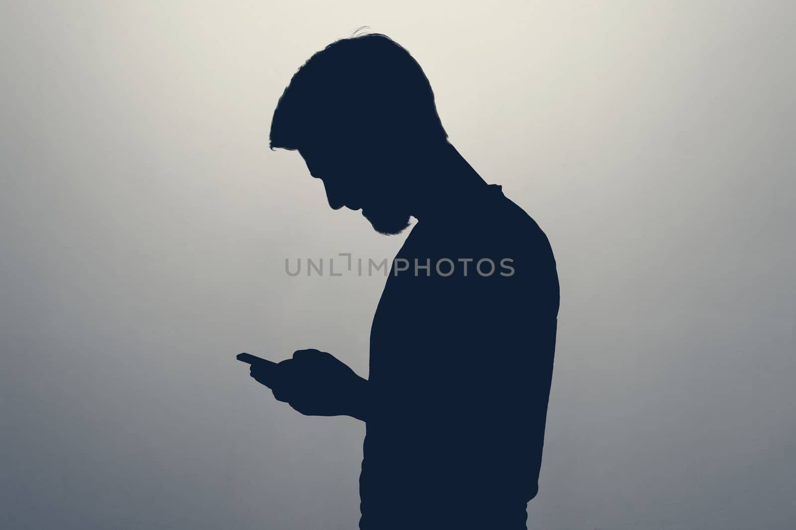 Male silhouette standing isolated on gray background looking at screen of smart phone, browsing web pages and chatting with friends.