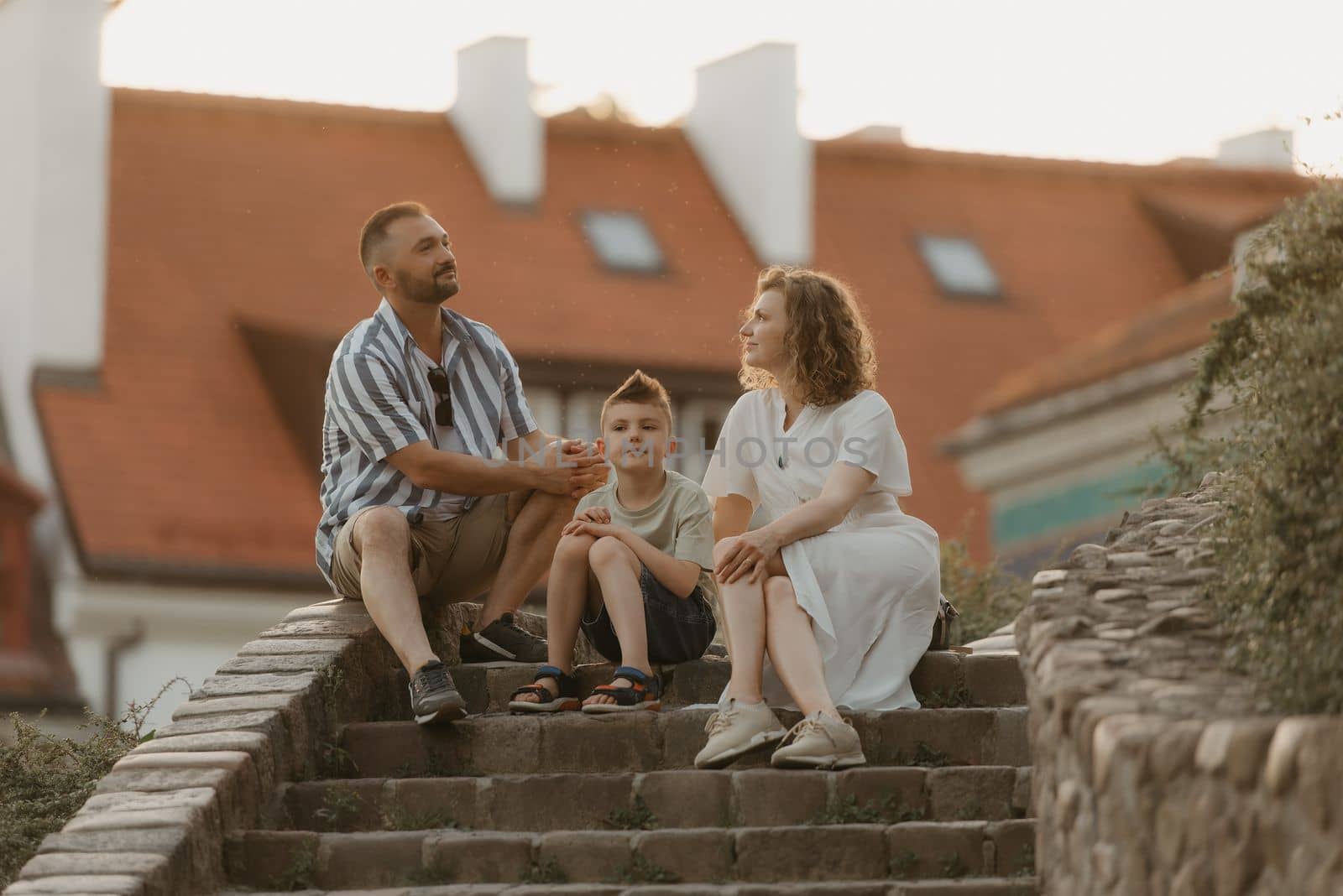 A family is sitting on the stairs between roofs in an old European town. A happy father, mother, and son are having fun in the evening.