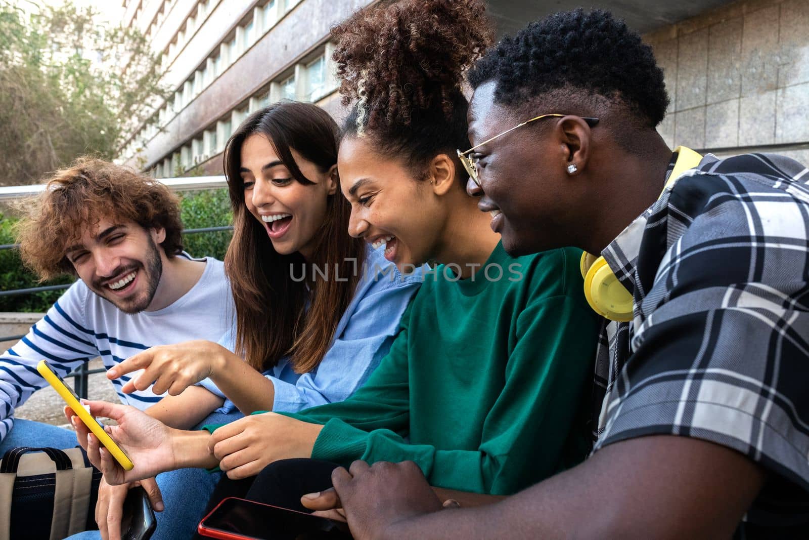 Multiracial college student friends look at mobile phone laughing together. Group of happy people using smartphone outdoors. Youth lifestyle and social media addiction concept.