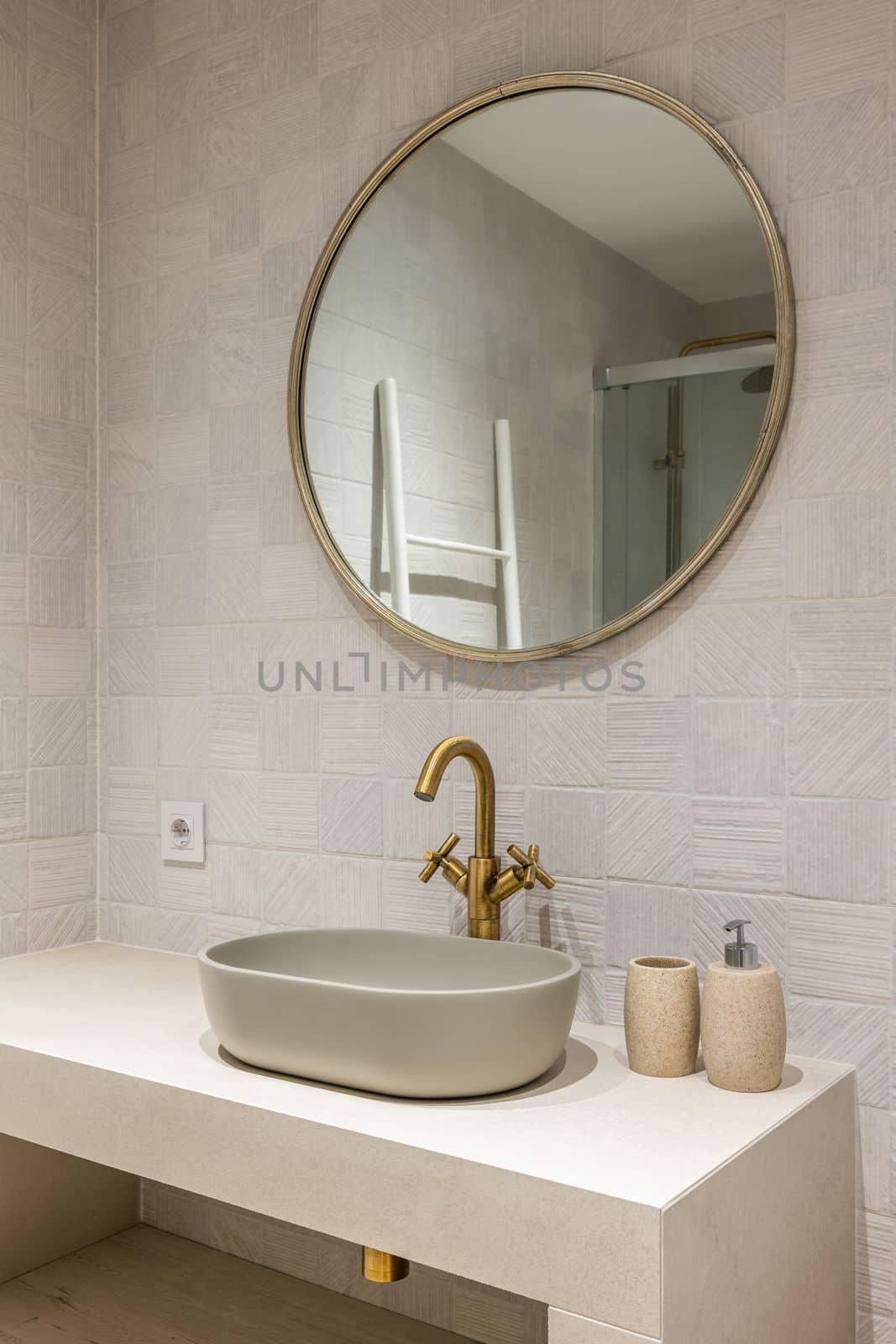 Neat stylish oval-shaped sink on white marble countertop with an elegant copper-coloured faucet. Above sink hangs a round modern gold-framed mirror that reflects part of shower. by apavlin