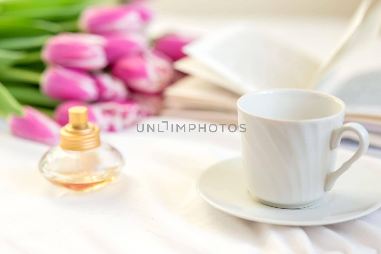 A still life with a white mug and a saucer, a golden bottle of perfume, an open book and a bouquet of beautiful pink tulips lies on a bed on a white fabric. close-up. copy space