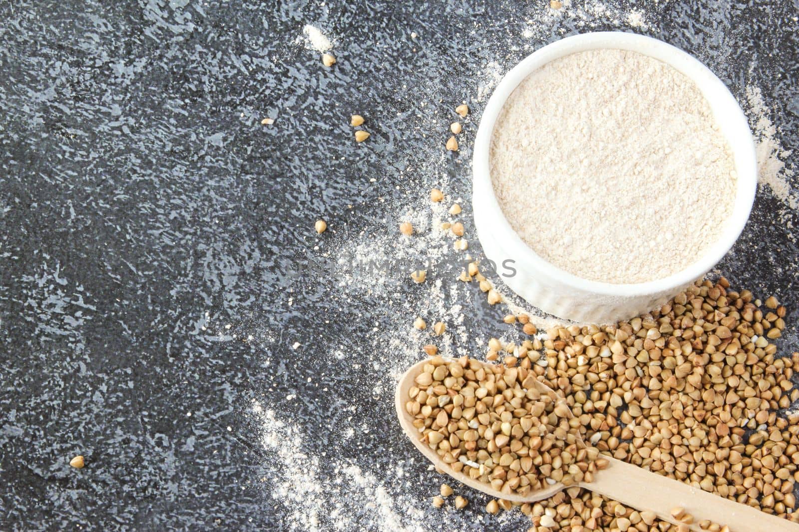 Buckwheat flour in a white bowl and wooden spoon, raw green buckwheat grain on concrete stone background, close up, top view. Alternative flour, gluten free flour, healthy nutrition