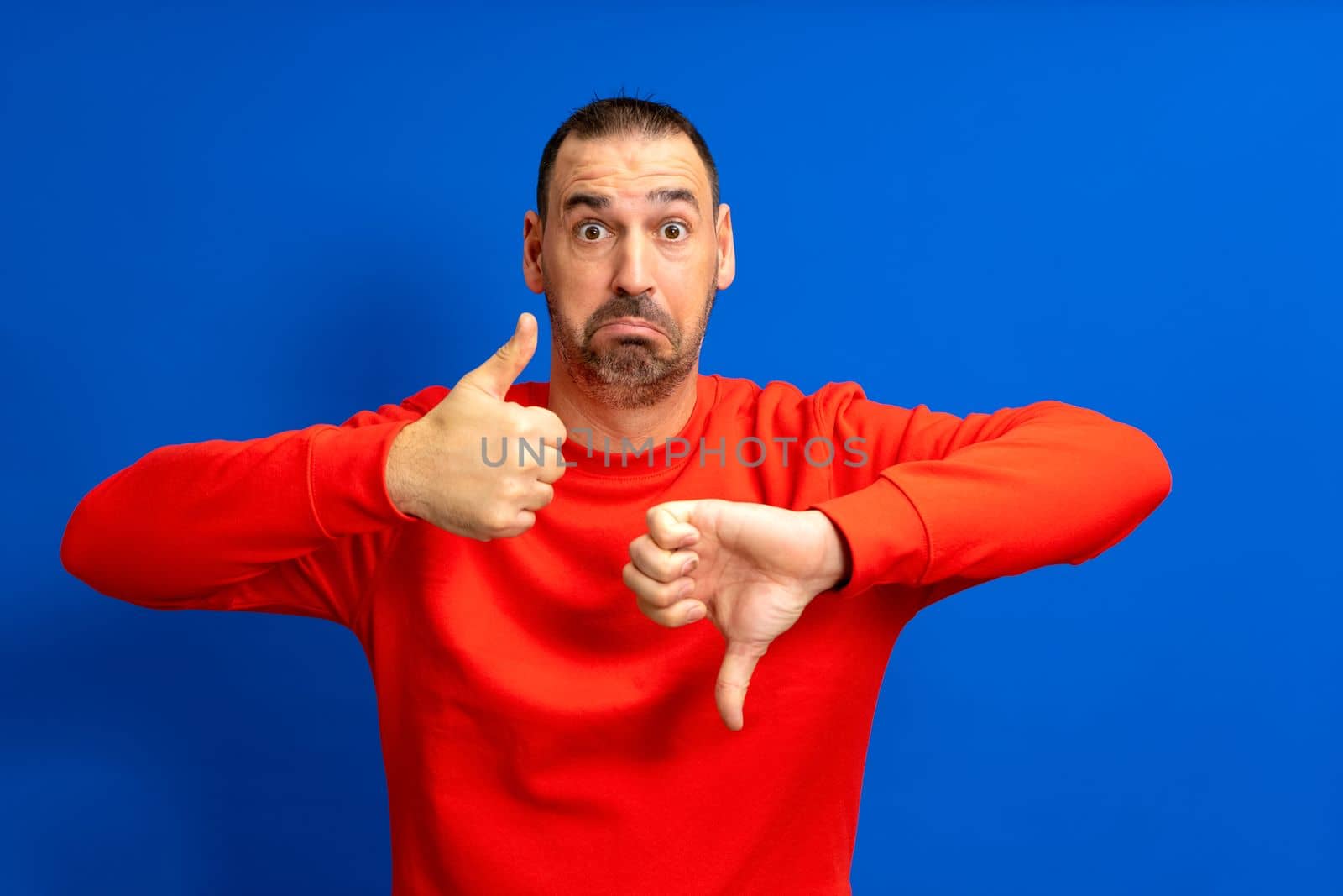 Photo of doubtful handsome bearded latino man wearing red jumper showing thumb up and down over isolated blue color background