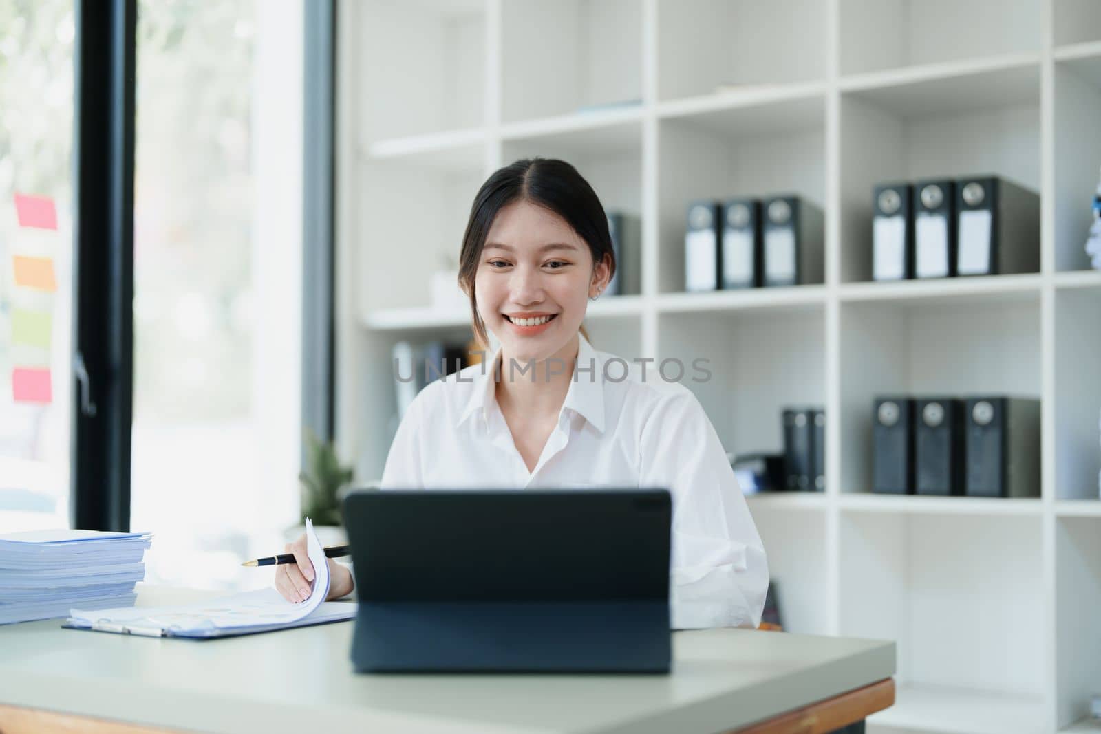 Portrait of a thoughtful Asian businesswoman looking at financial statements and making marketing plans using a computer on her desk by Manastrong