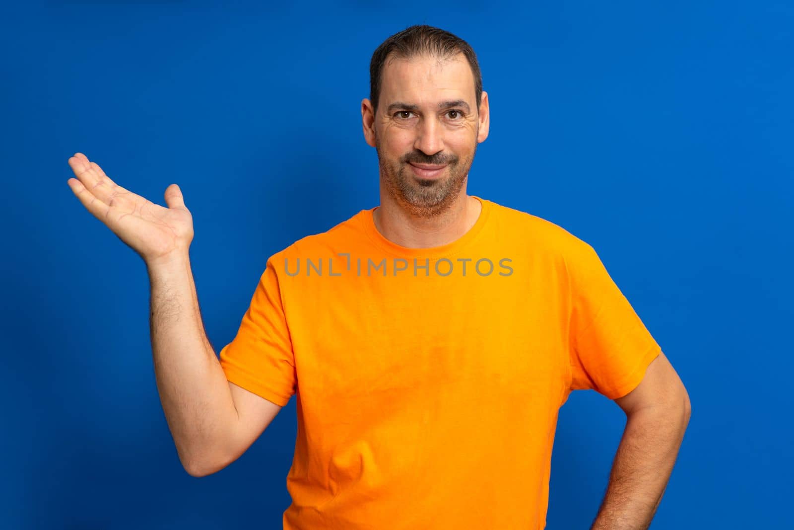 Handsome latino man offering something imaginary in the palm of his hand with friendly and affable attitude, isolated on blue studio background