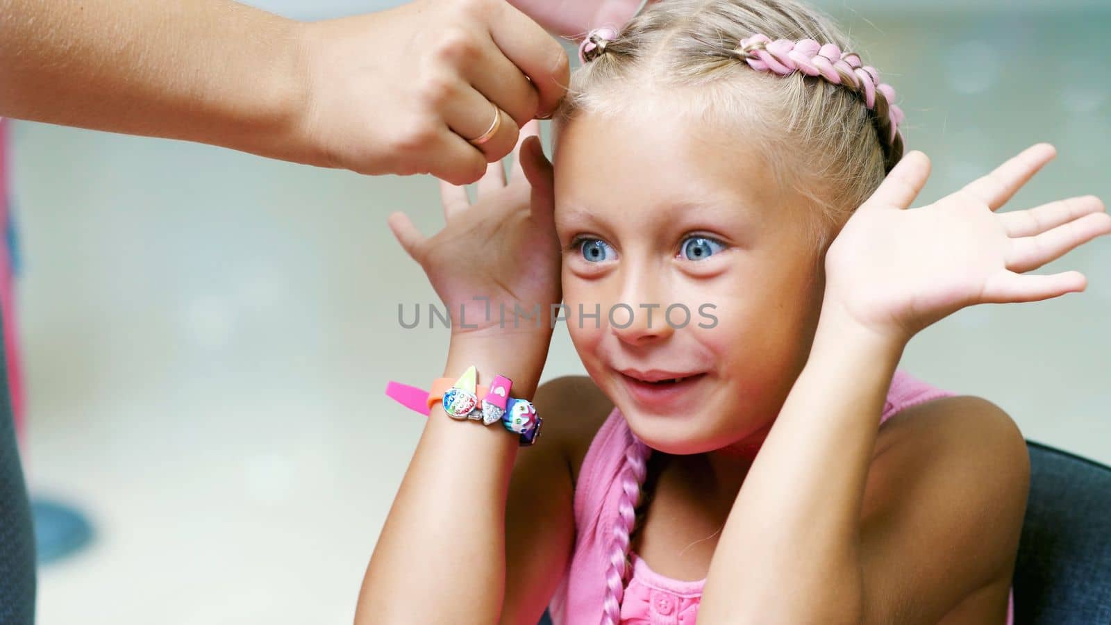 Beautiful blond girl, of seven years old, braided two pigtails, do a hairstyle with pink locks of hair in a beauty salon, a hairdresser's salon, in front of a large mirror. a little princess. High quality photo