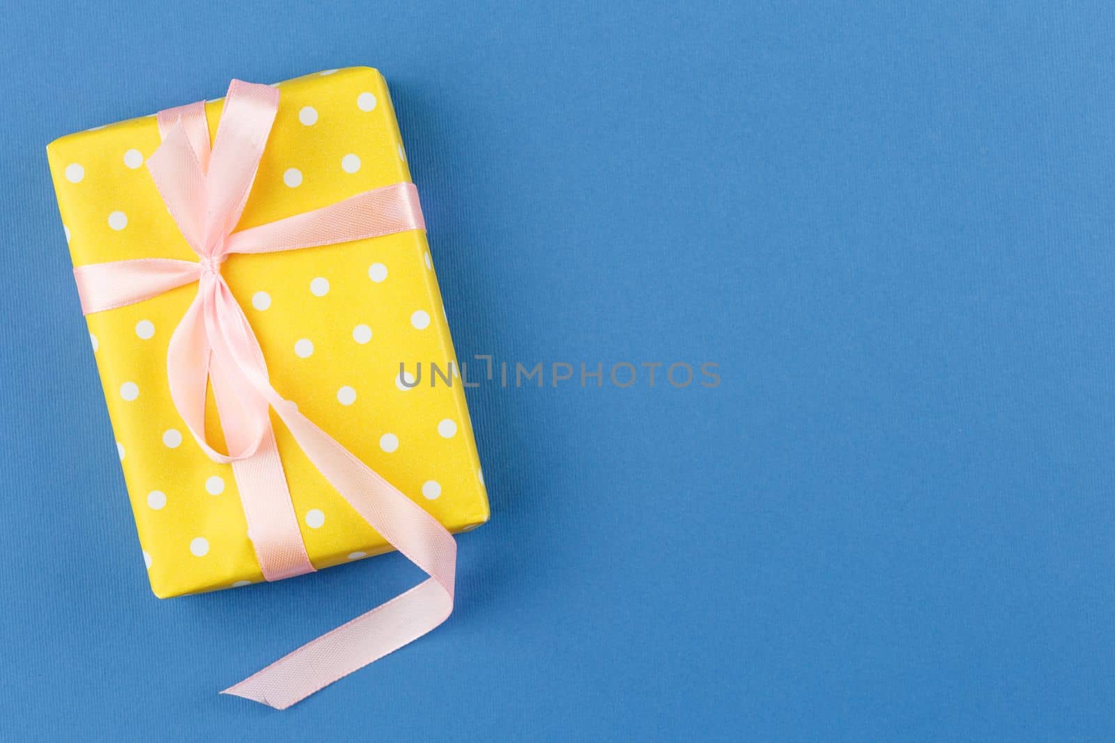 Gift in yellow wrapping paper with polka dots on blue isolated background. Flat lay. Present box with satin pink bow. Top view.