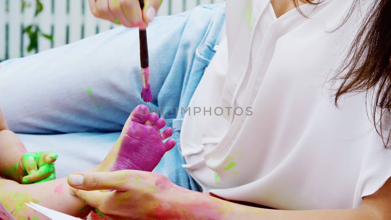 a young beautiful mother and her one-year-old daughter playing with finger paints in the garden, sitting on a plaid, a blanket, a coverlet, on the grass, lawn, in the summer. High quality photo