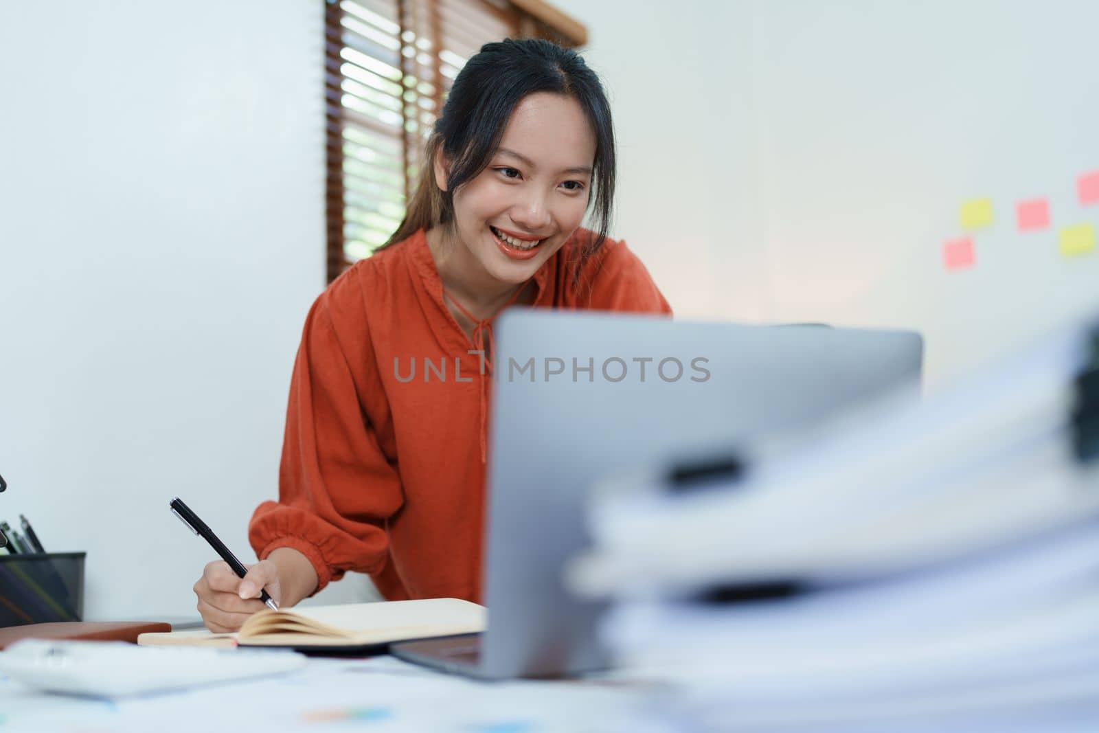 Portrait of a thoughtful Asian businesswoman looking at financial statements and making marketing plans using a computer and notebook on her desk by Manastrong