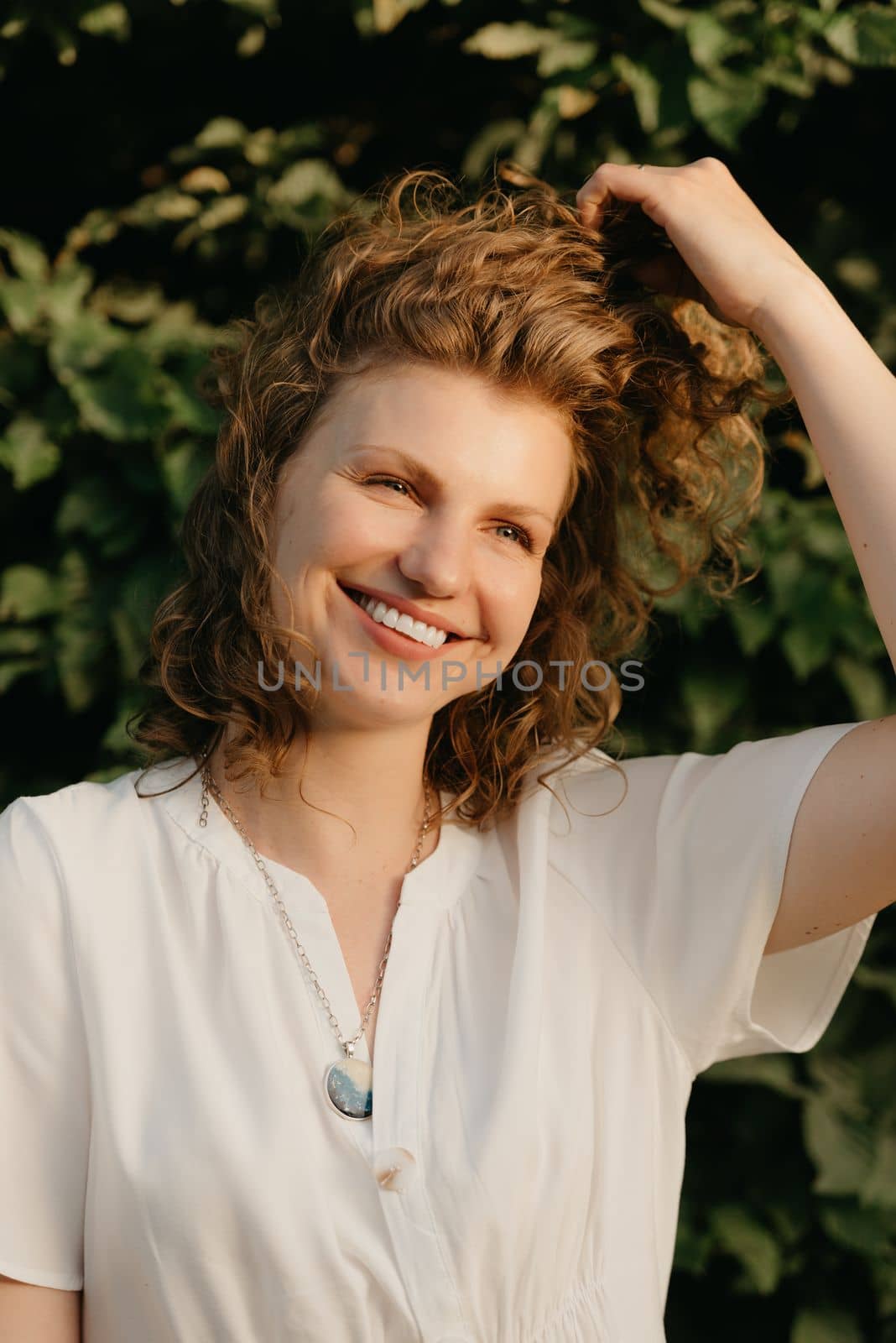 A smiling woman with curly hair is posing in the park in the evening. A happy lady in a white dress with her arm on her head with green leaves in the background.