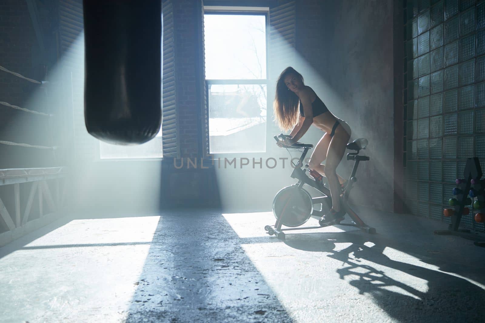 The beautiful fitness girl with a sport body twists bicycle pedals in gym, large loft-style room, beautiful light from windows, sun beams, the stationary exercise machine the bicycle by vladimirdrozdin