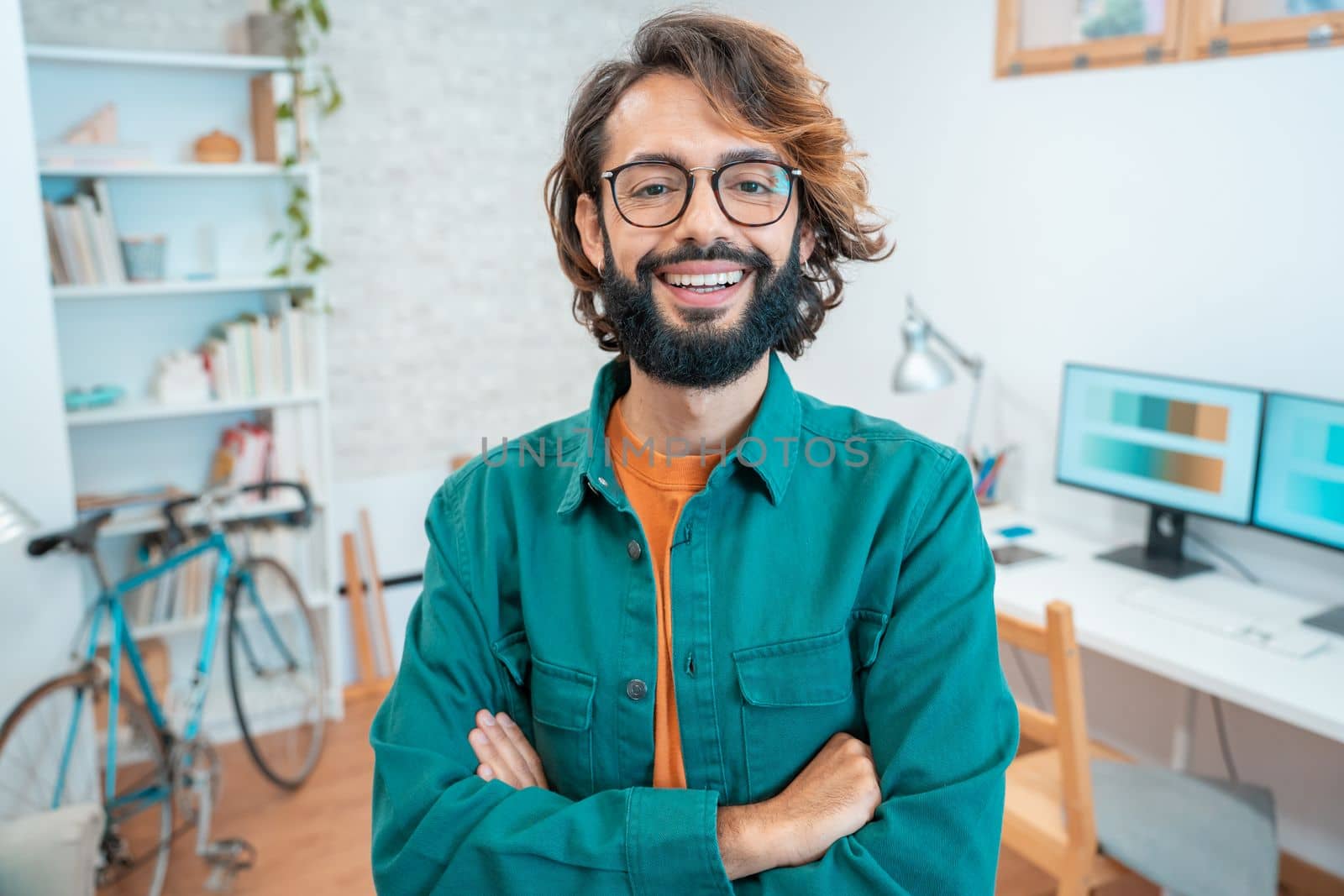 Portrait of a young creative entrepreneur smiling looking to camera in modern workplace - Portrait of millennial freelancer man in home office with bike, glasses and green jacket. High quality photo