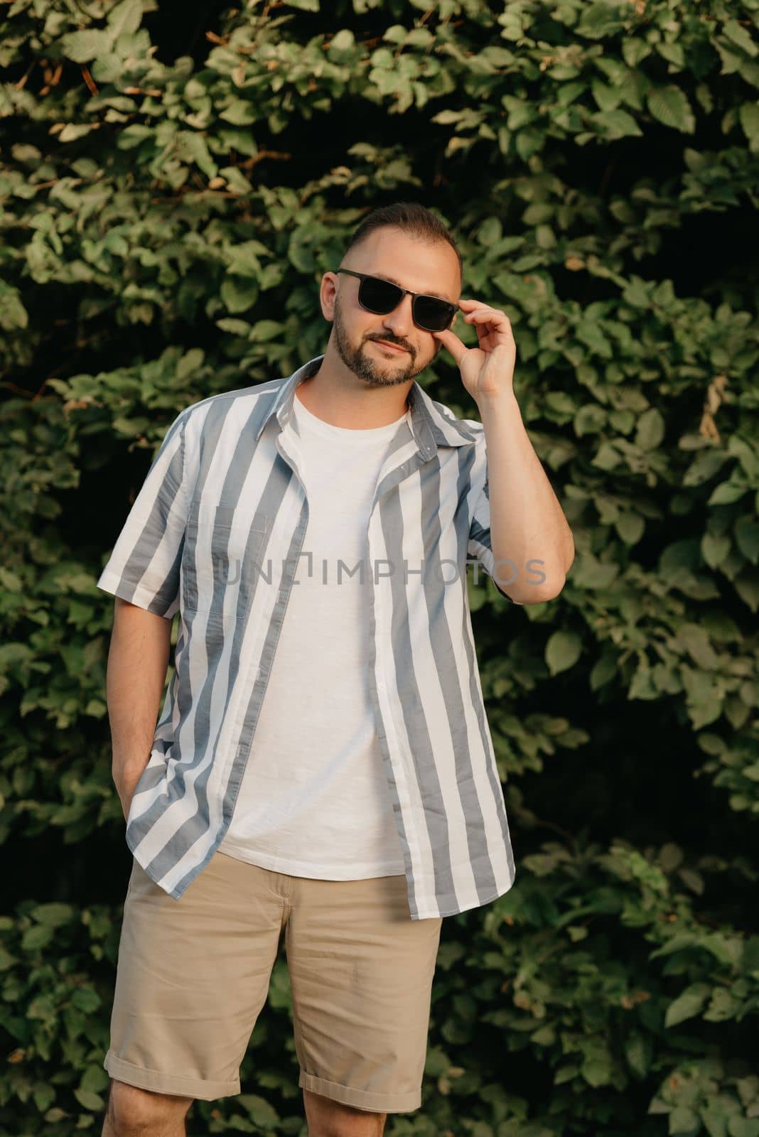 A smiling man is posing in the garden in the evening. A happy guy is touching his sunglasses with green leaves in the background.