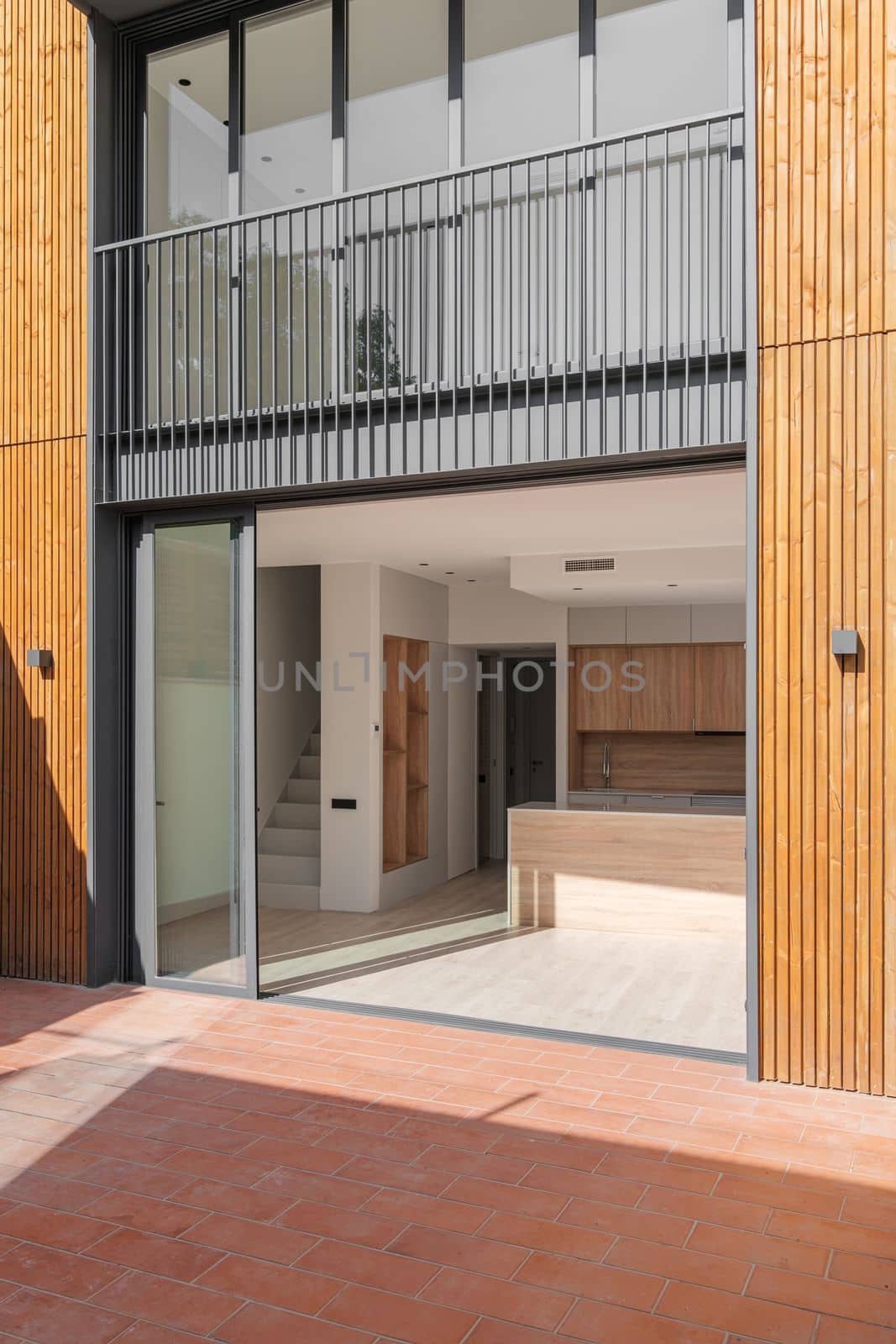 Glass doors in black metal frame open onto house with spacious kitchen with light wooden furniture and staircase to second floor. Doors connect the back of the house and the patio