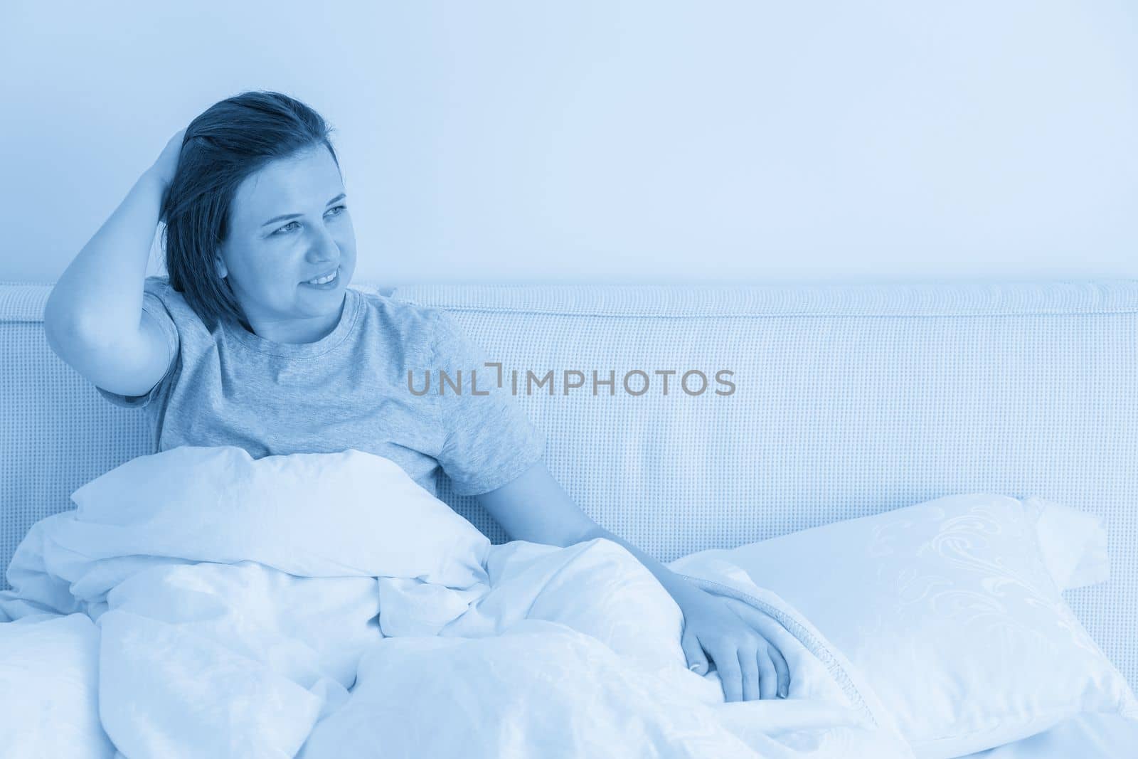 Smiling brunette woman wearing pajama sitting in bed in bedroom by Mariakray