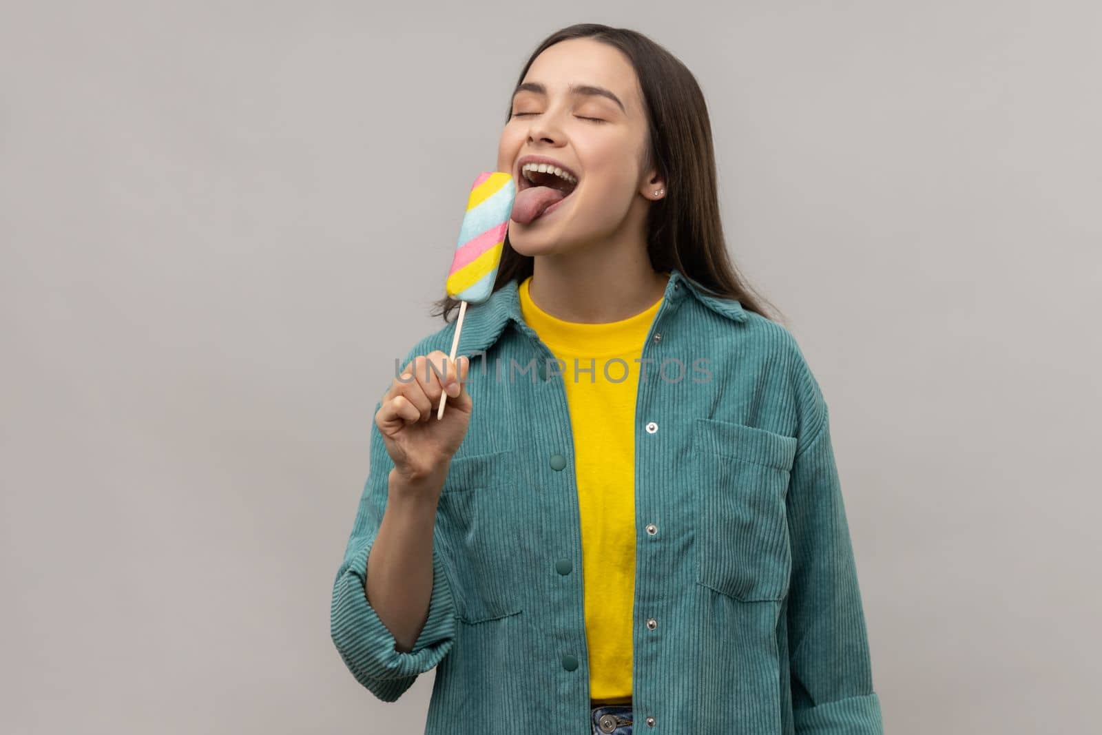 Portrait of positive woman with dark hair licking multicolored ice cream, feels hungry, tasting dessert, wearing casual style jacket. Indoor studio shot isolated on gray background.