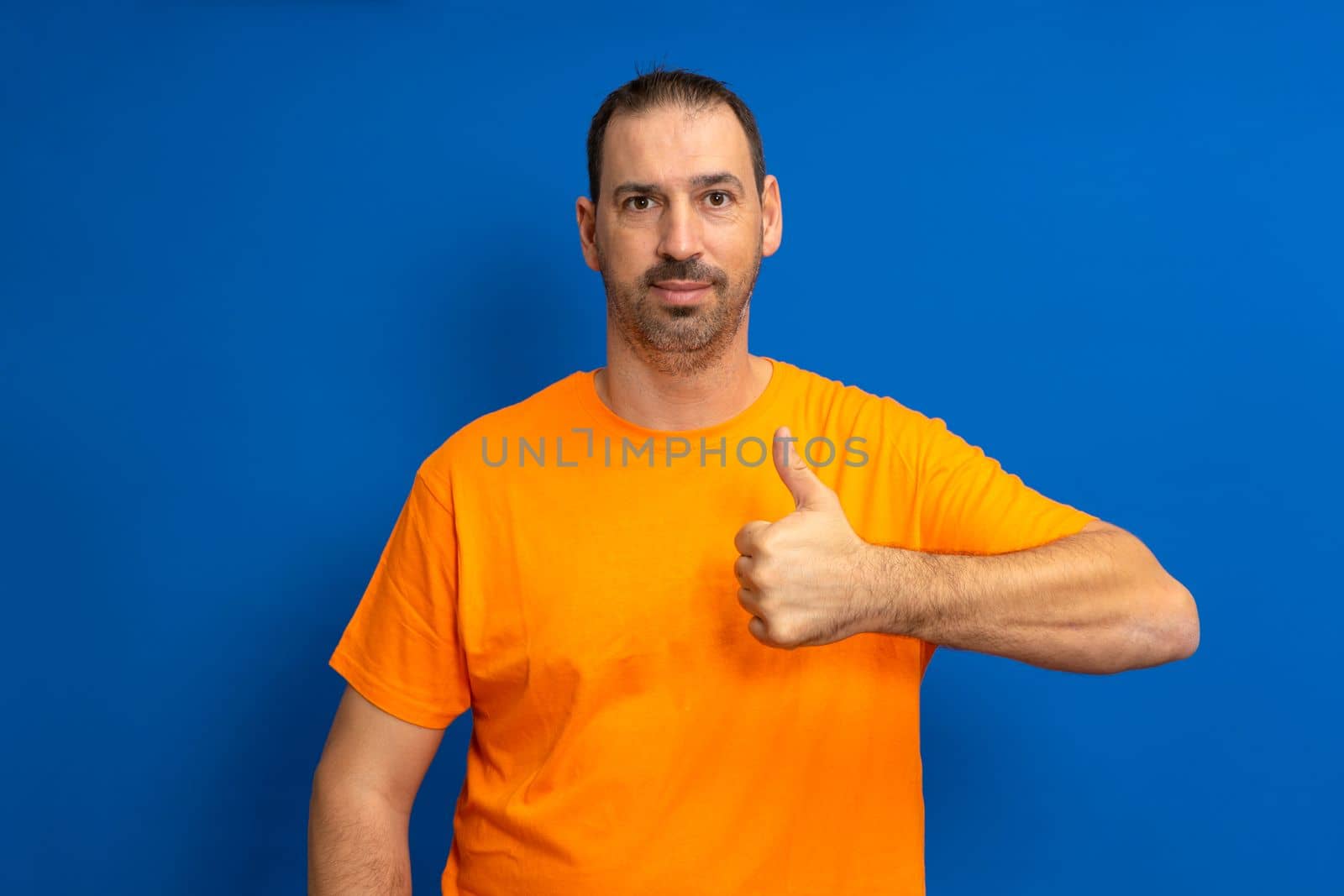 Bearded handsome hispanic man wearing orange casual t-shirt standing over isolated blue background doing happy thumbs up gesture with hand. Approval expression looking at camera showing success.