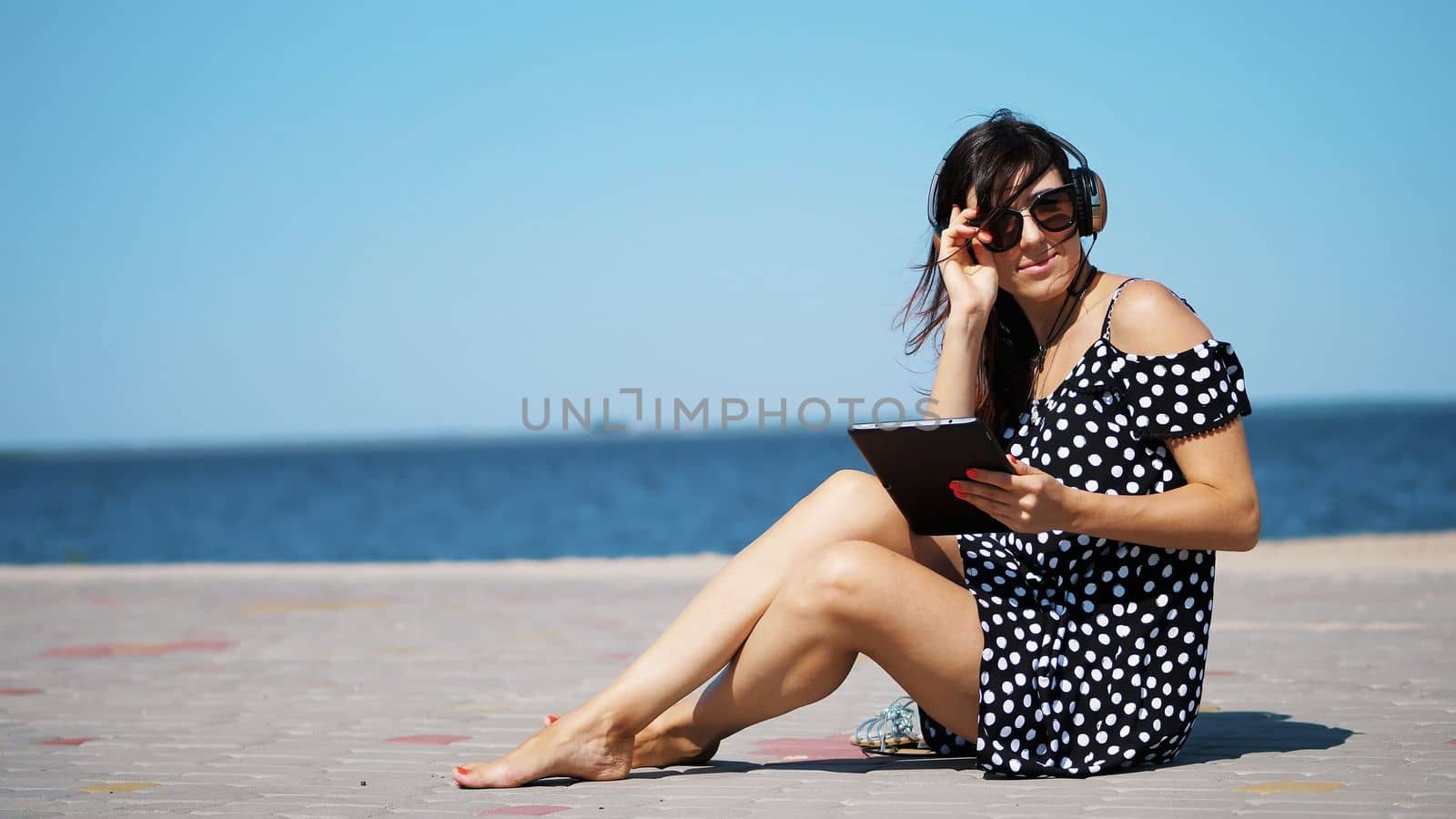 beautiful brunette, woman in sun glasses, and headphones, works on a tablet, listening to music , sitting on the beach, barefoot, against the sea. wind waves her hair. High quality photo