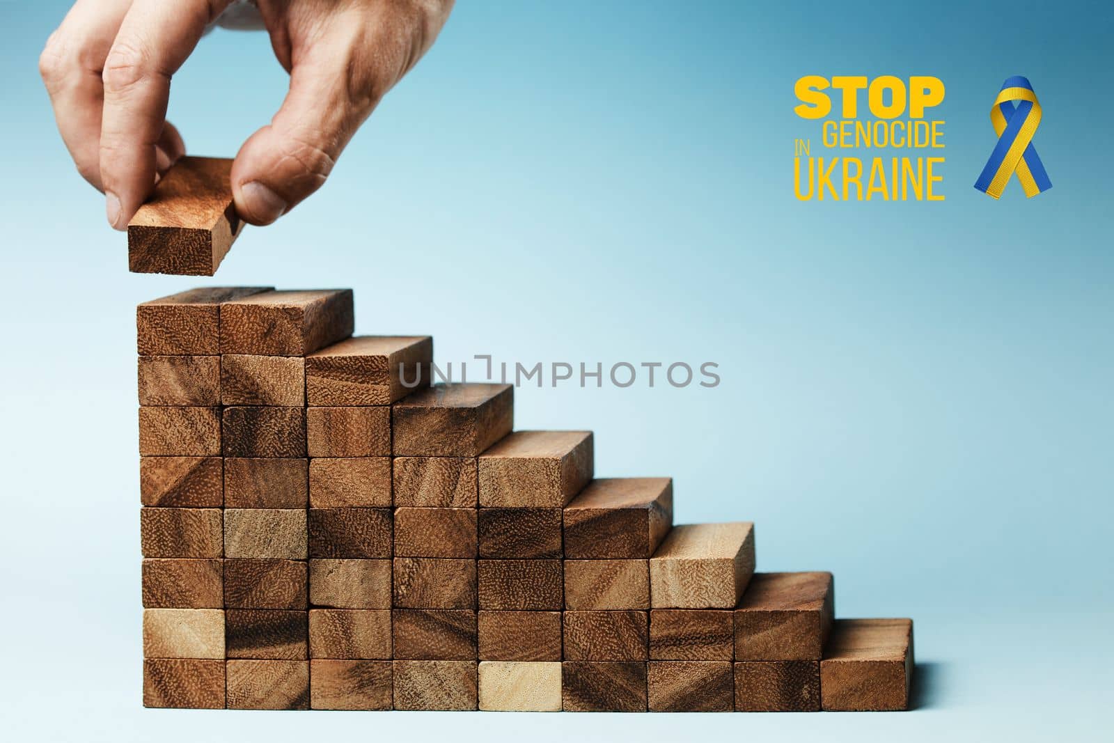 hand lays the last detail on ladder made of wood, on blue background with words stop genocide in ukraine. concept needs help and support, truth will win
