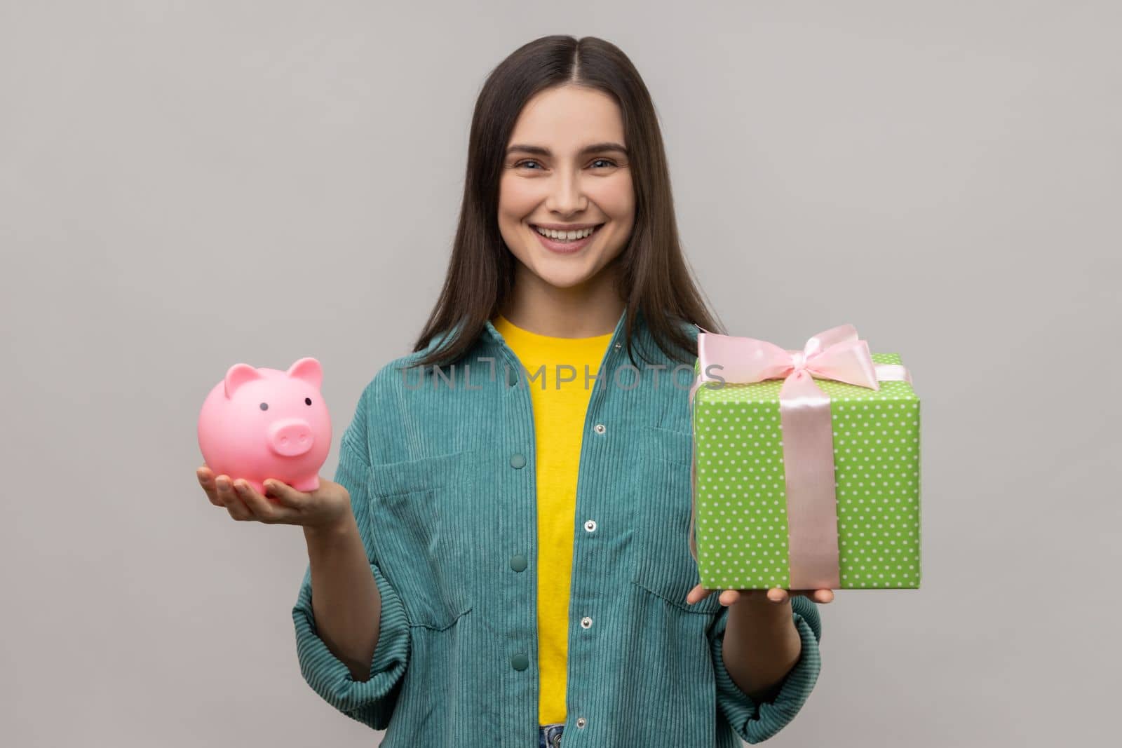 Portrait of happy woman holding wrapped gift box and piggy bank, save money, advertising, looking smiling at camera, wearing casual style jacket. Indoor studio shot isolated on gray background.