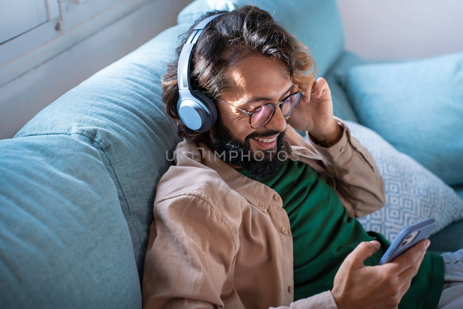 Smiling young man wearing headphones listen music playing in smartphone sitting on sofa at home by PaulCarr