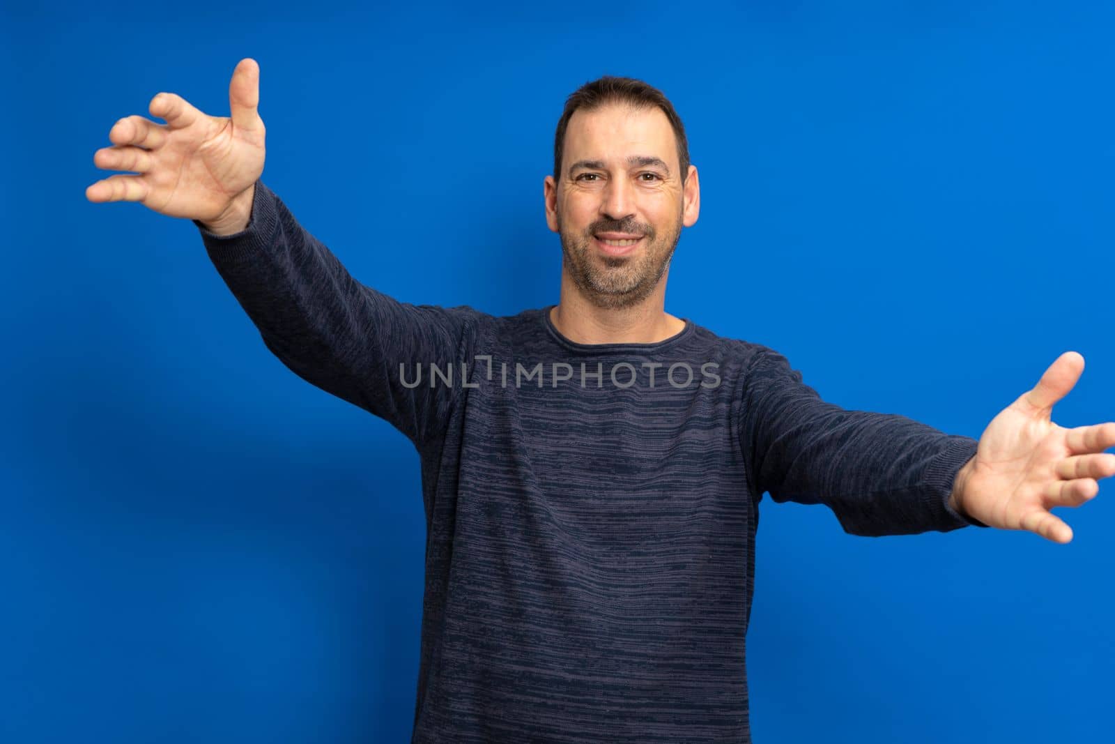 Happy handsome bearded hispanic man standing with arms raised, wants to hug his friend, isolated on blue background... I love you. I miss you body language