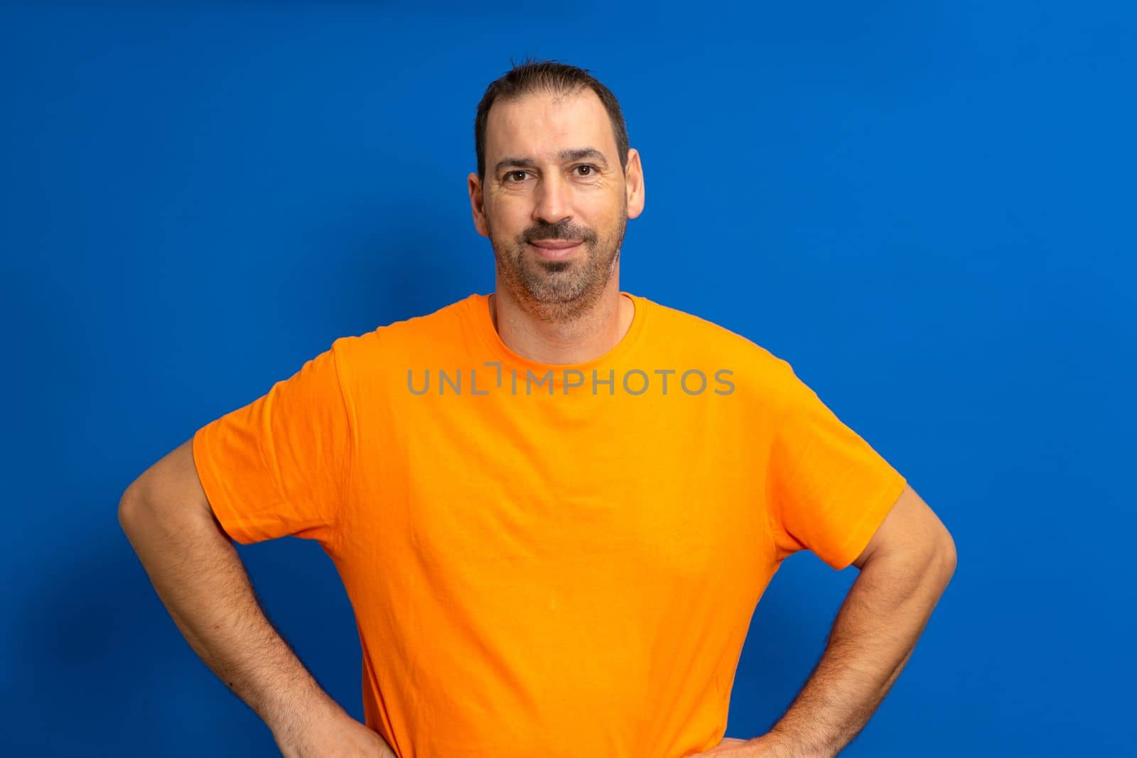 Cheerful bearded man in orange t-shirt posing isolated on blue background, studio portrait. People lifestyle concept. Mock up copy space. Standing with arms akimbo on waist blinking
