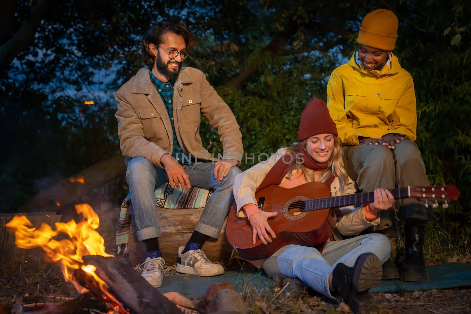 Three happy diverse friends having fun playing music and enjoying bonfire camping in nature at night by PaulCarr