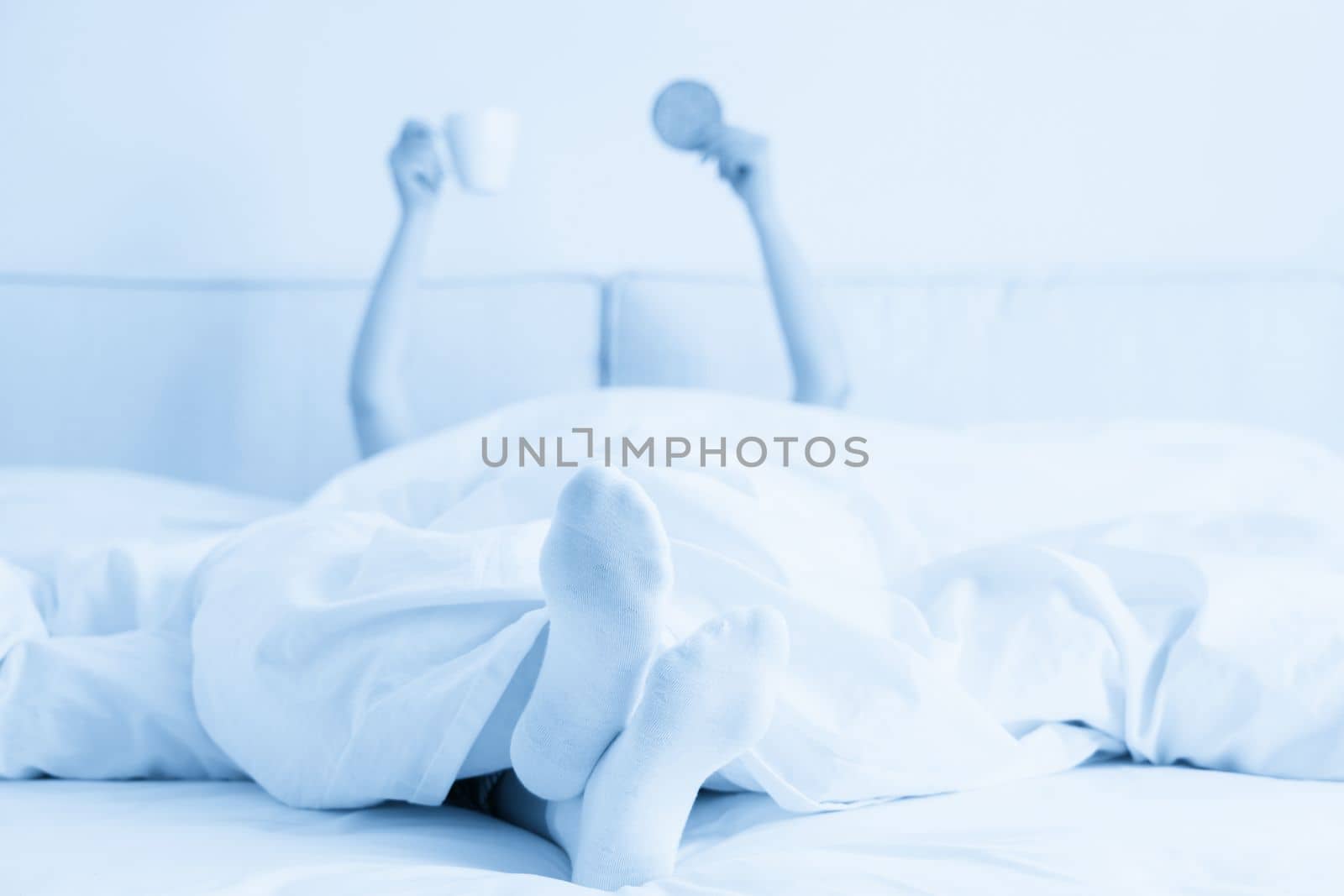 Female hand holding cup of coffee and cracker from under a blanket in bed. Woman waking up in the morning by Mariakray
