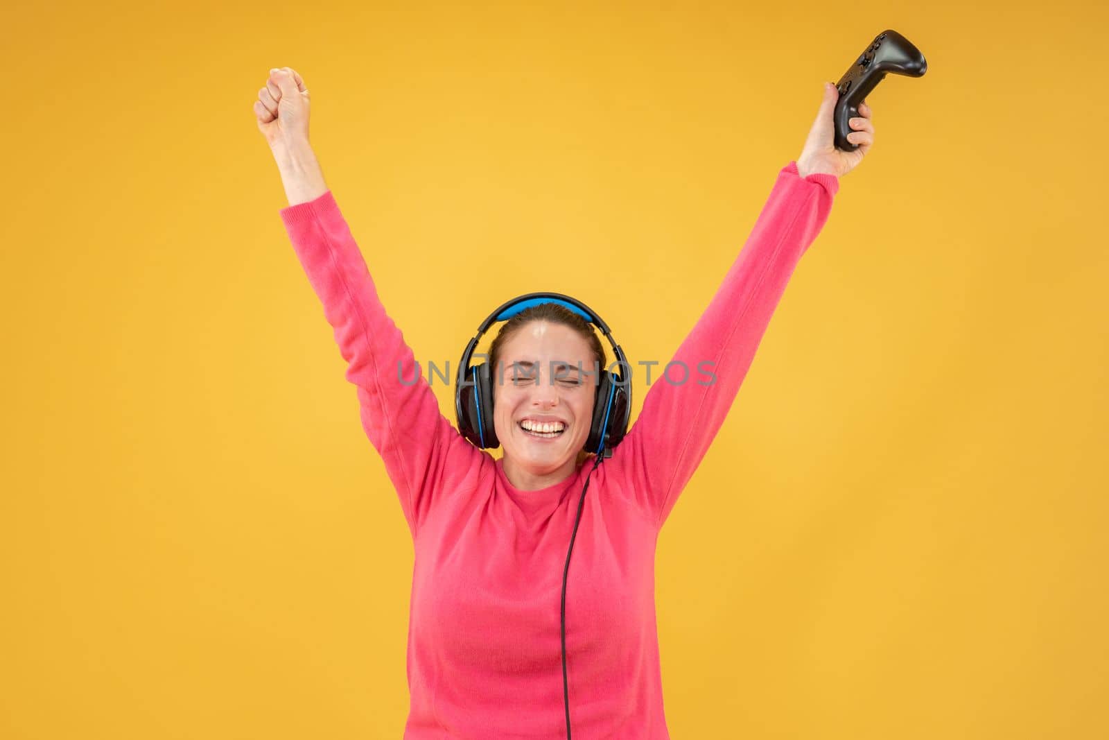 Beautiful woman playing games wearing headphones standing isolated yellow background. Excited smiling female with raised arms celebrating victory. Success concept. High quality photo