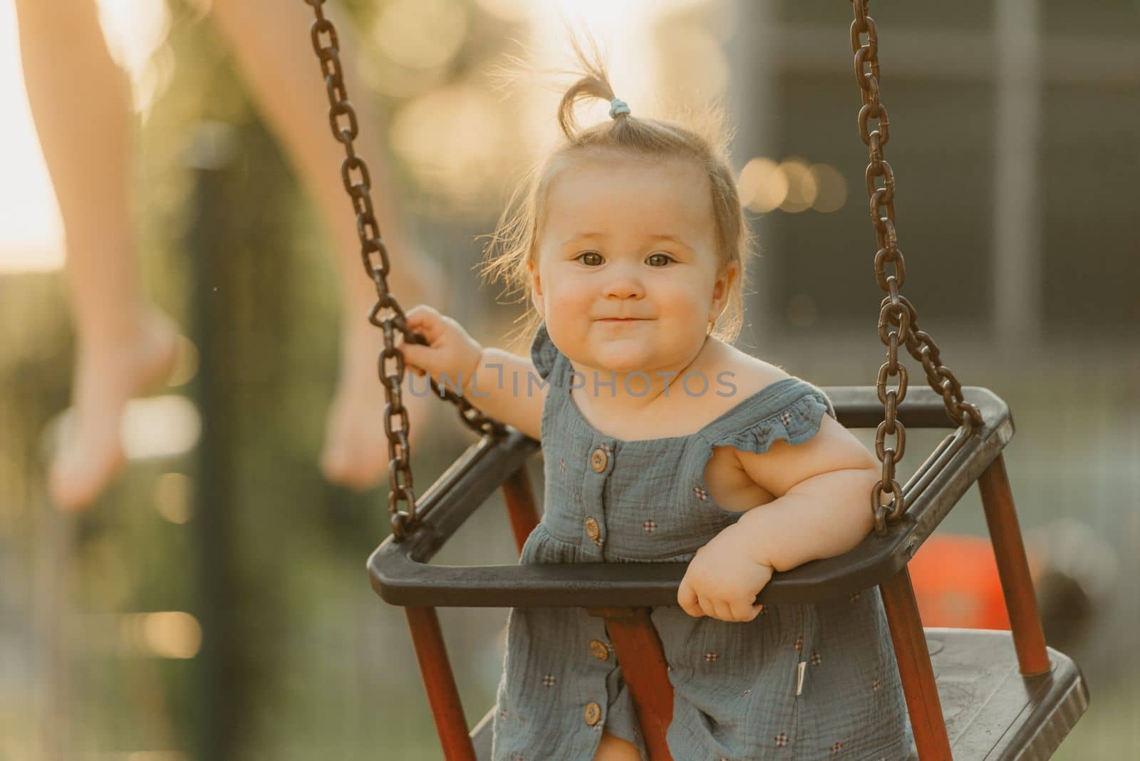 Toddler baby girl on a swing on the warm summer evening. Mother is swinging her young daughter on a sunny playground.