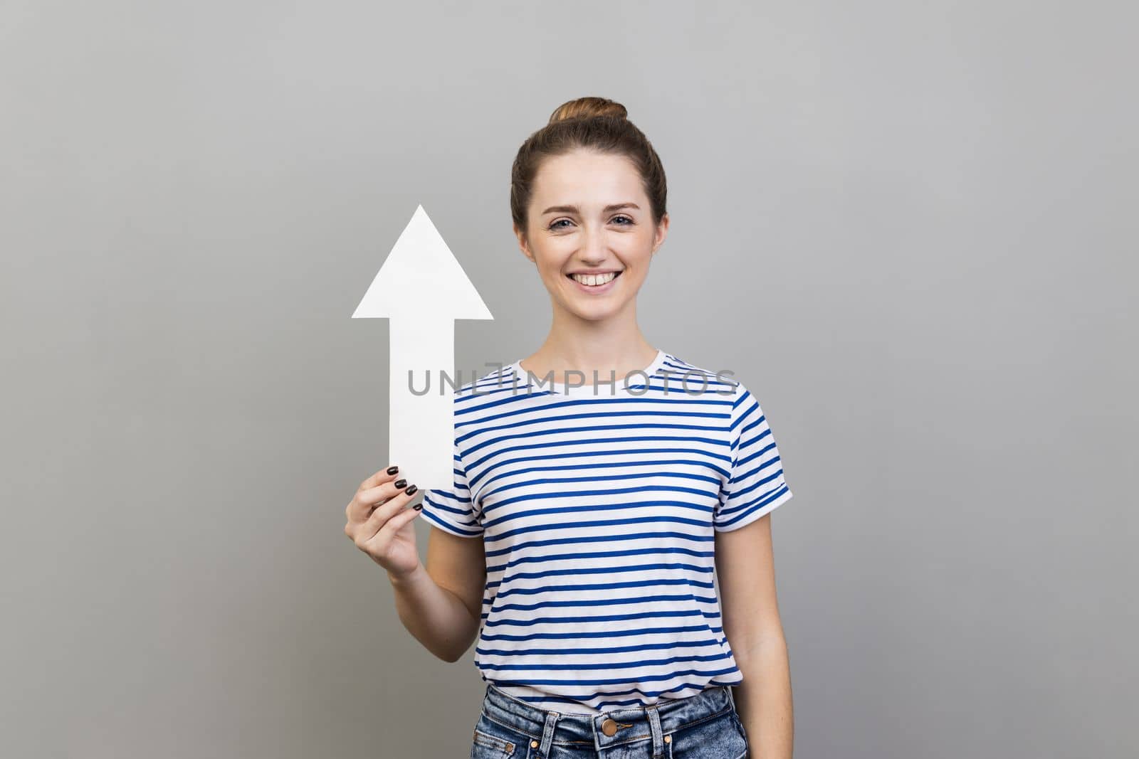 Portrait of woman wearing striped T-shirt holding arrow pointing up looking at camera with smile, growth and increase concept. Indoor studio shot isolated on gray background.