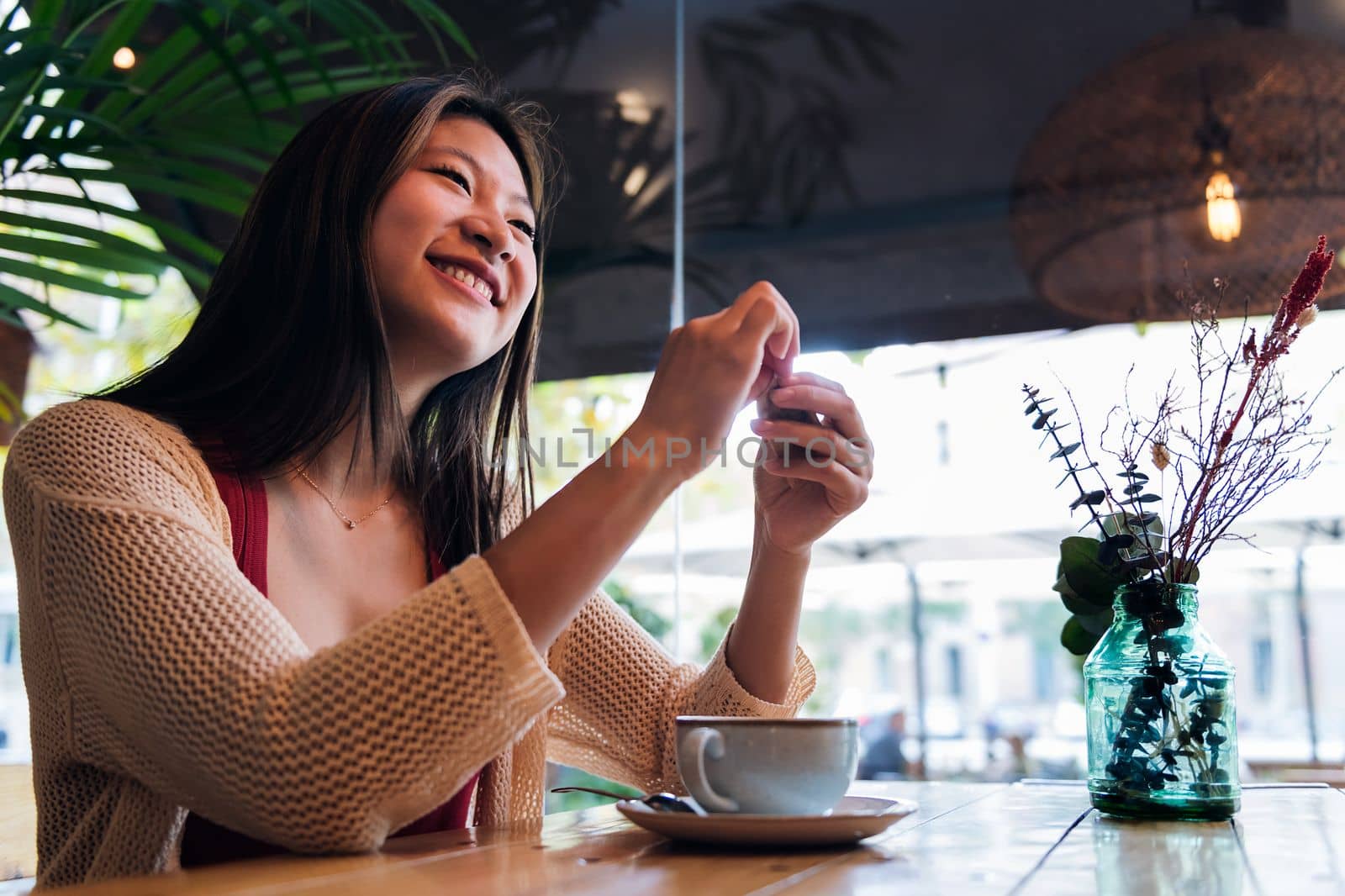 smiling asian woman sitting in a coffee shop opening the sugar sachet for her coffee, concept of relaxation and leisure