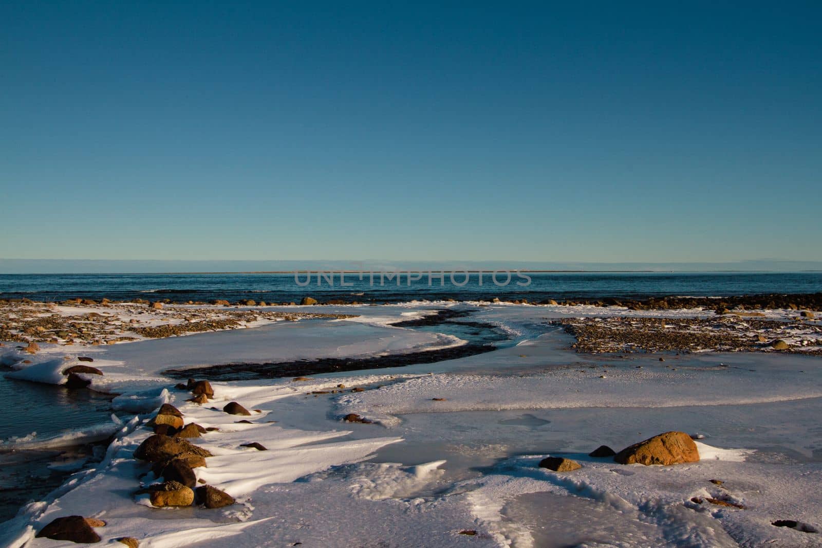 Arctic landscape - a small creek flowing into a larger waterbody surrounded by frozen arctic tundra with snow covered rocks on a clear cold day, near Arviat, Nunavut