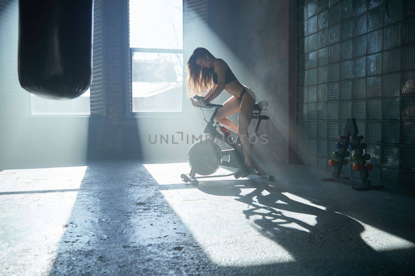 The beautiful fitness girl with a sport body twists bicycle pedals in gym, large loft-style room, beautiful light from windows, sun beams, the stationary exercise machine the bicycle. High quality photo
