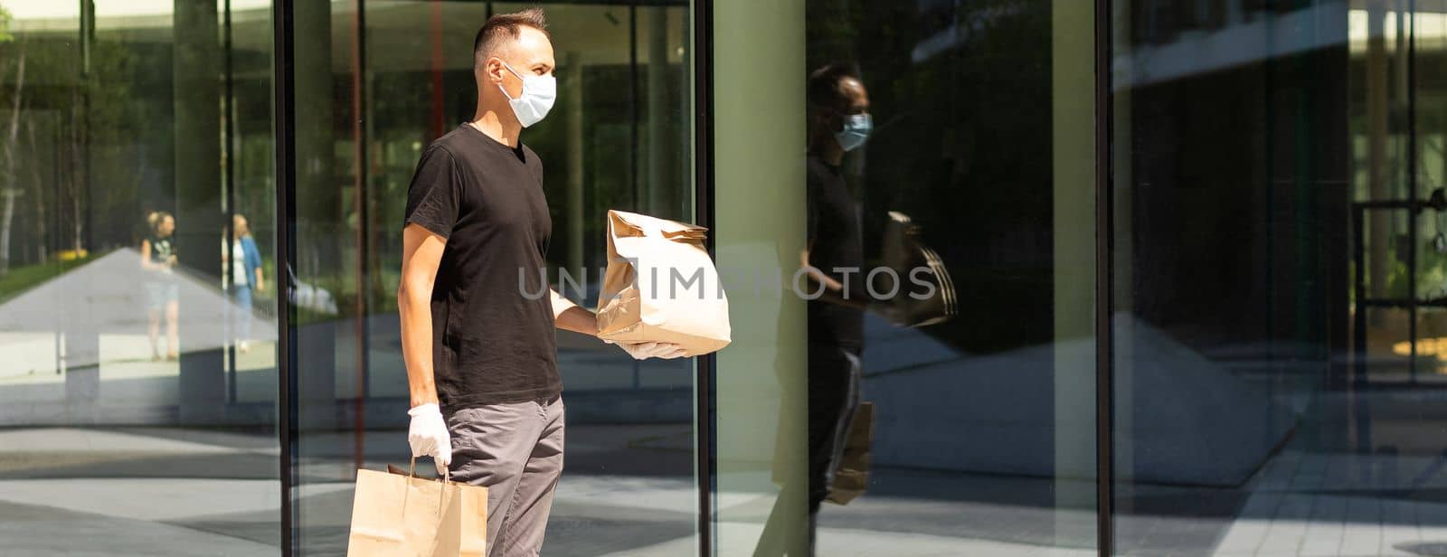 Delivery guy with protective mask and gloves holding bag with groceries in front of a building