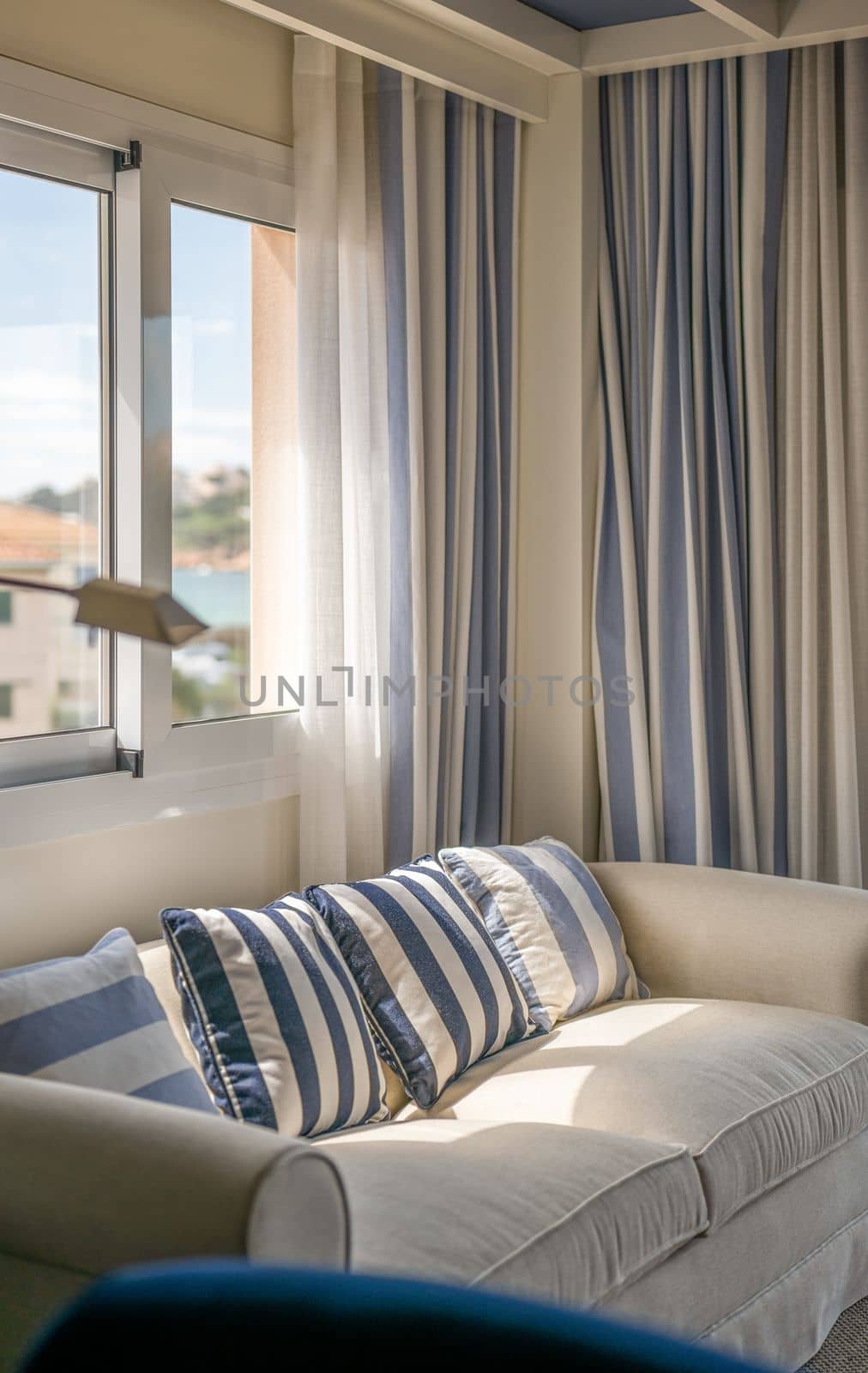 Closeup of cozy upholstered sofa in beige color with decorative pillows in vertical blue-beige stripes and curtains in same style. Cozy living room flooded with bright sunlight from window
