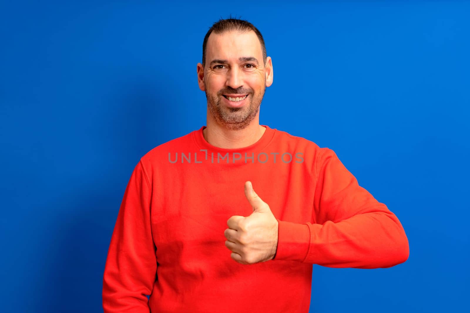 Bearded latin man wearing a red sweater raising his thumb up in approval while smiling at the camera in a friendly attitude isolated over blue background. by Barriolo82