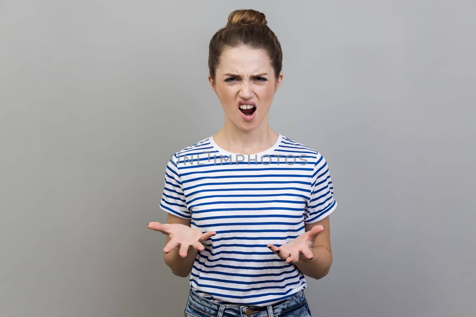 Portrait of angry unhappy frustrated woman wearing striped T-shirt standing spreading hands with shock and misunderstanding, how could you sign. Indoor studio shot isolated on gray background.