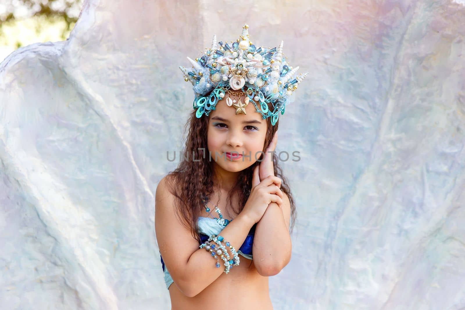 A beautiful little girl in a turquoise crown and mermaid costume is standing outdoors, in a large seashell. Close up. Copy space