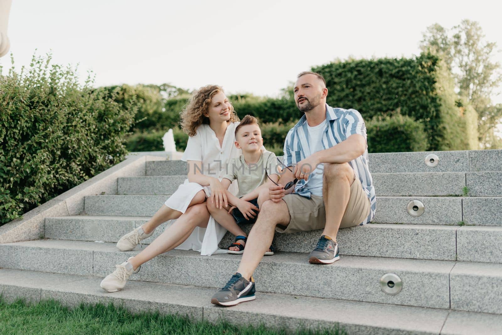 Father, mother, and son are sitting on the steps in the garden of an old European town. Happy family in the evening. Dad is discussing important themes with his smiling family in the park at sunset.