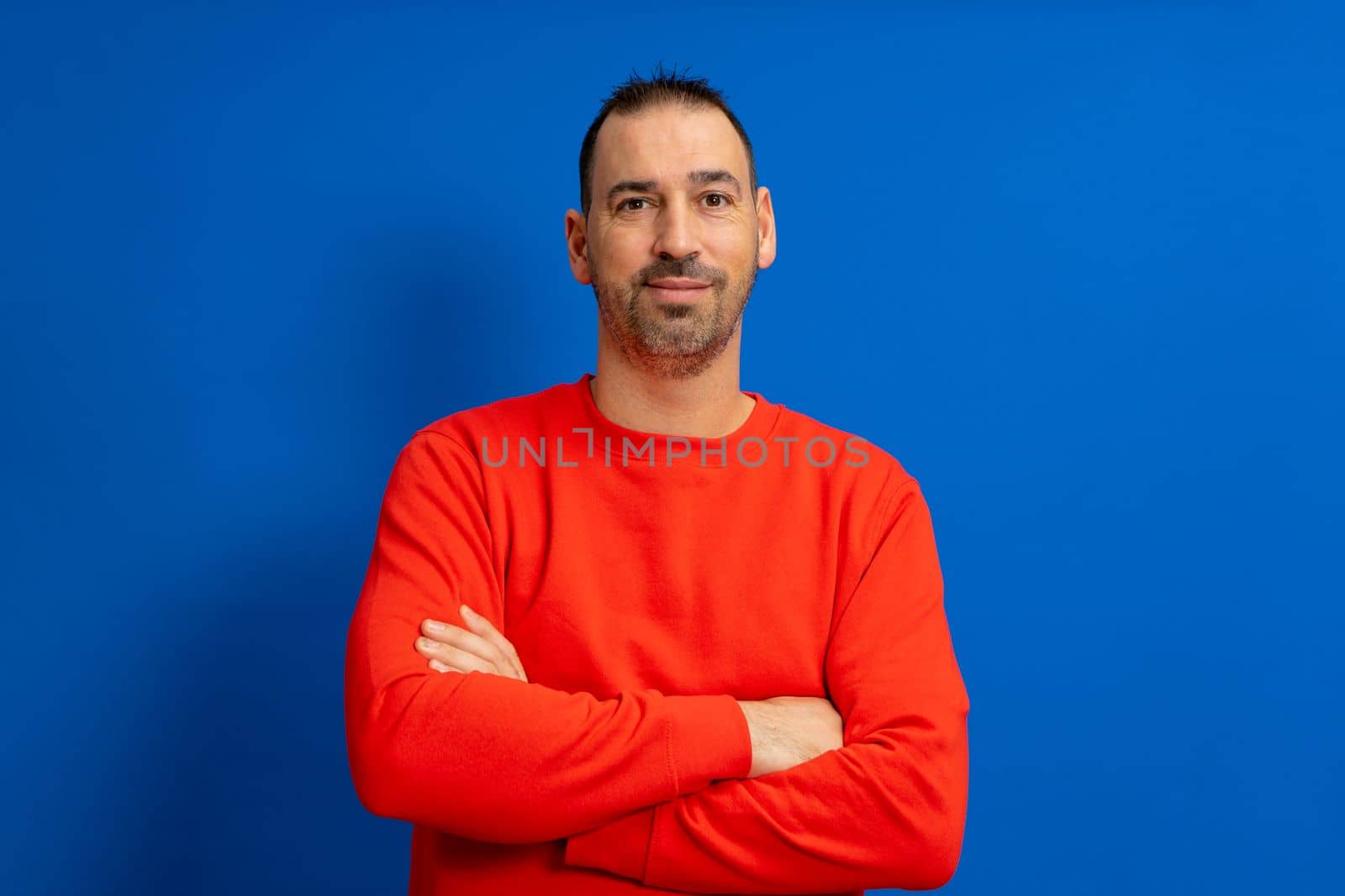 Portrait of smart cheerful attractive bearded hispanic man with arms crossed wearing bright red pullover isolated over blue colored background