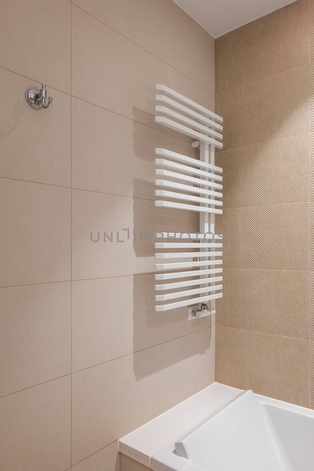 Corner of bathroom in the center with a white heating radiator on the wall for drying towels. The walls of the room are lined with beige tiles of different colors. Pure white acrylic bathtub. by apavlin