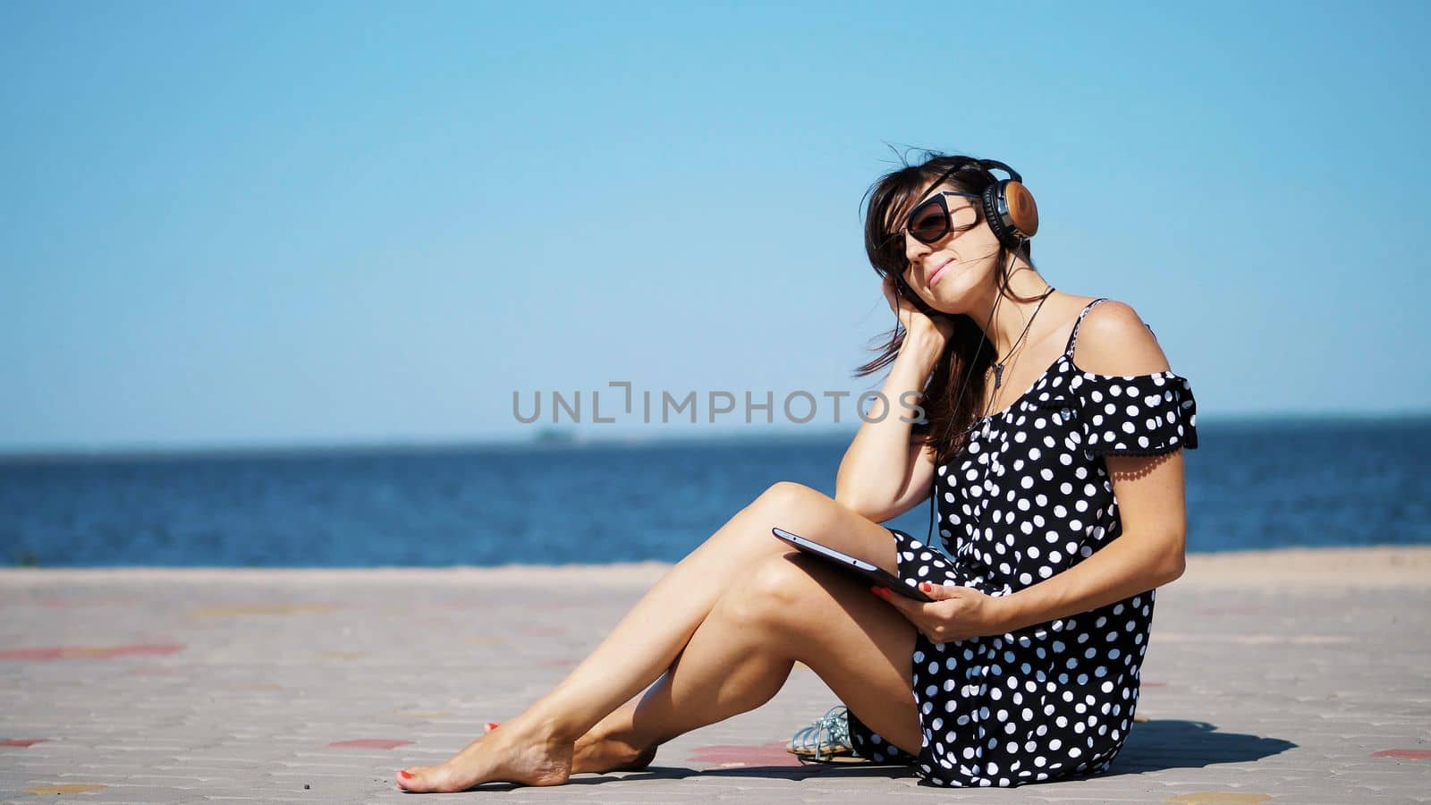 beautiful brunette, woman in sun glasses, and headphones, works on a tablet, listening to music , sitting on the beach, barefoot, against the sea. wind waves her hair. High quality photo